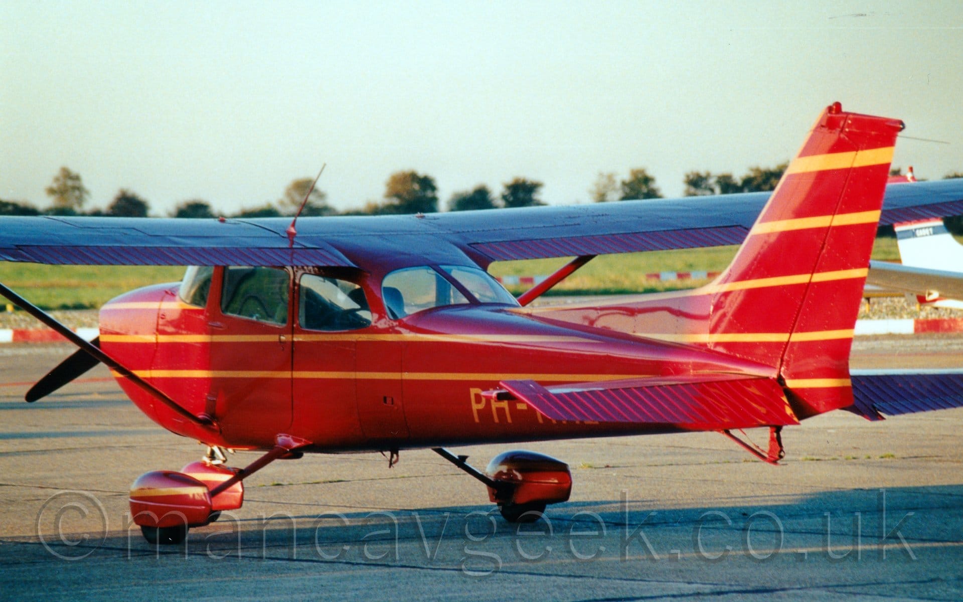 Side view of a red, high-winged, single propellor-engined light aircraft parked facing to the left and slightly away from the camera. The plane has thin yellow stripes running along the body and tail. The registration is on the lower rear fuselage in yellow, only the first part, "PH-" is visible, the rest hidden by the horizontal stabiliser. The plane is casting a deep shadow on the ground in the foreground, while the ground in the background is brightly lit by bright sunshine. Trees line the skyline, under bright but hazy skies.