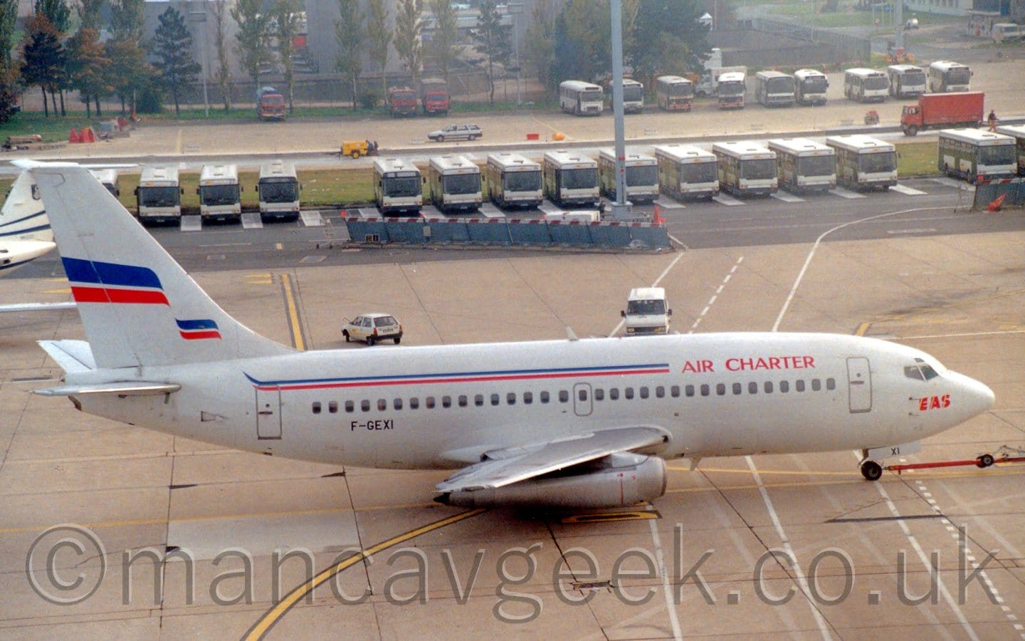 Side view of a white, twin engined jet airliner being towed from left to right by an out-of-frame truck, attached to the nose wheel by a long red pole. There is a thin blue and red stripe running along the top of the fuselage, and red "Air Charter" titles on the upper forward fuselage. There are additional red "EAS" titles on the nose. A wavy red and blue ribbon stretches across the tail. A row of white and blue buses parked side by side stretches across the frame, with trees and more buses further back.