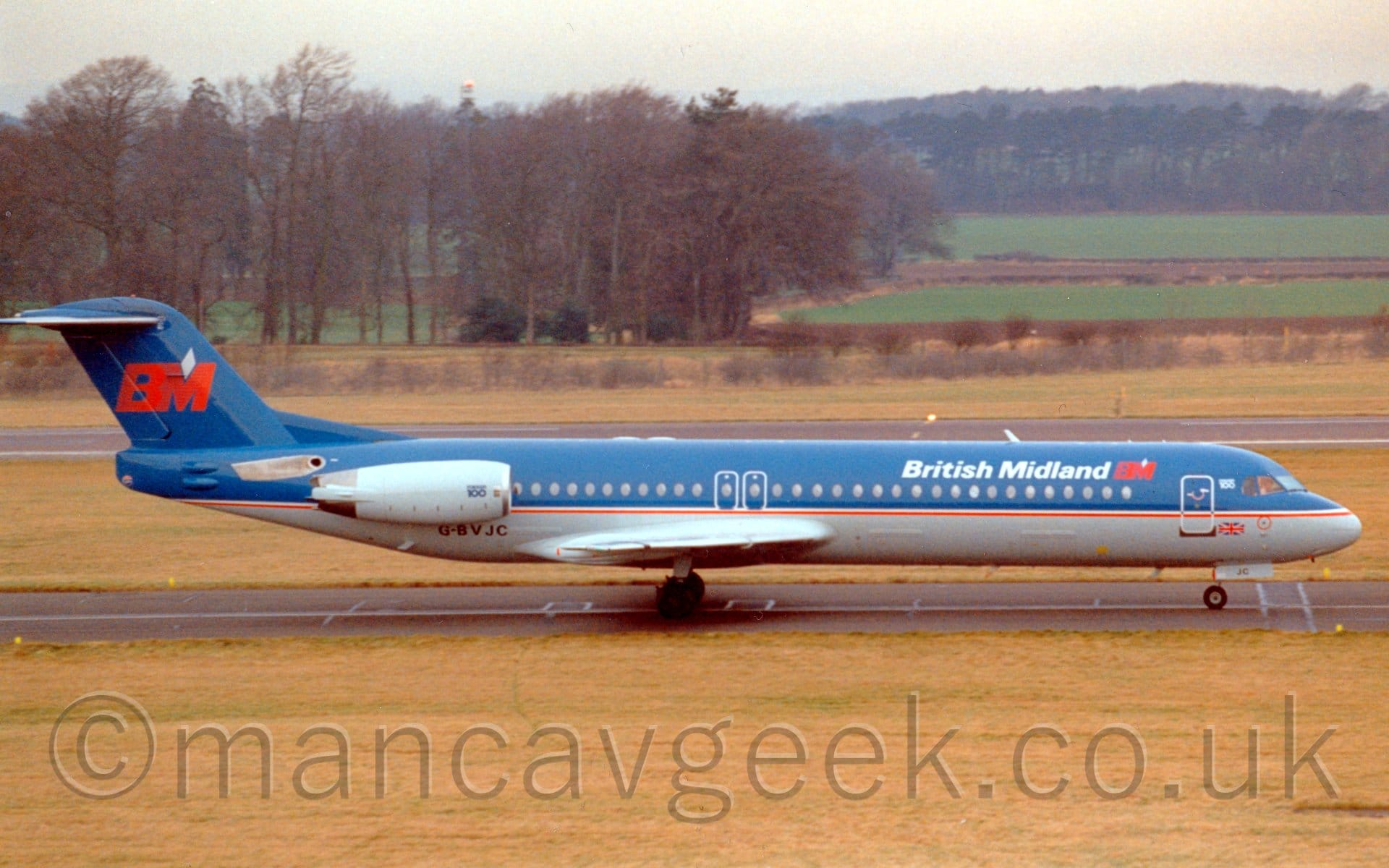 Side view of a blue, twin engined jet airliner taxiing from left to right. The engines are mounted on the sides of the rear fuselage. The plane has a grey belly, a red stripe running along the side of the fuselage, and white "British Midland" titles on the upper forward fuselage. There is a large red "BM" logo on the blue tail, with a smaller version on the upper forward fuselage just aft of the cockpit. A large grassed are fills the foreground, with more grass leading up to trees in the distance in the background, under grey skies.