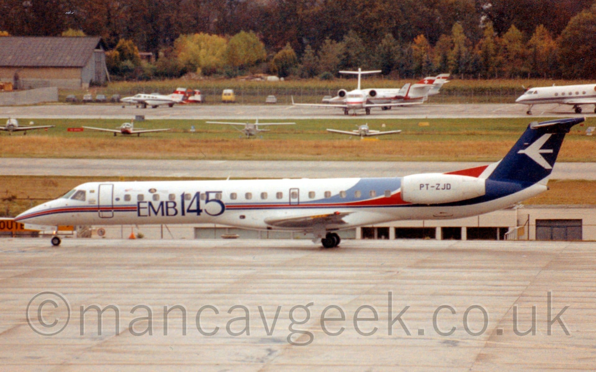 Side view of a long, thin, twin engined jet airliner with the engines mounted on the sides of the rear fuselage, taxiing from right to left. The plane has a thin light blue, red, and dark blue stripe running along the body, getting wider as it sweeps up towards the rear fuselage and tail. There are large blue "EMB145" titles on the forward fuselage, and the registration "PT-ZJD" in black on the engine pods. Grey concrete apron fills the foreground, with a handful of light aircraft parked on grass in the background, leading up to another apron area filled with BizProps and BizJets, with trees beyond that.