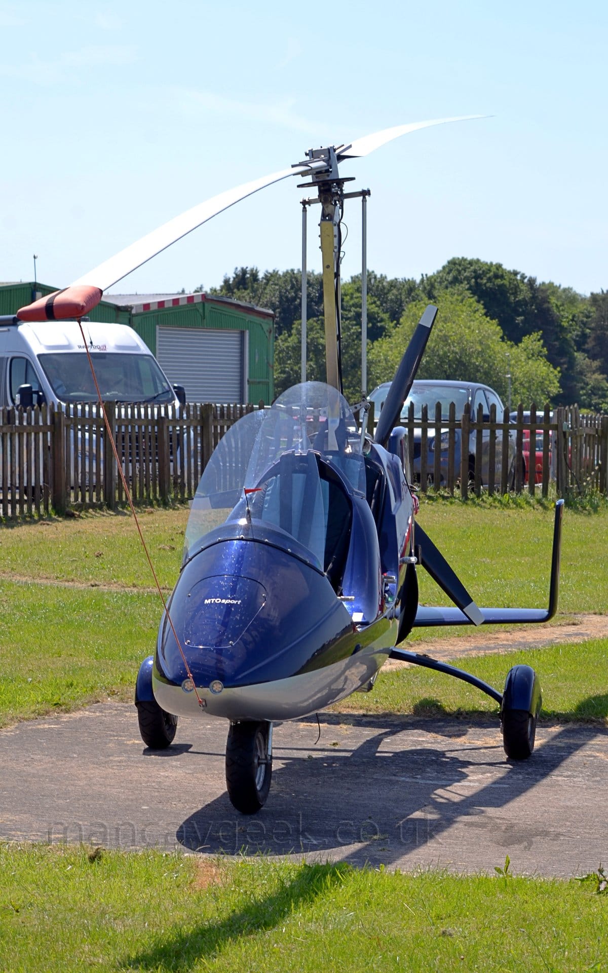 Front view of a blue, 2 seat gyrocopter parked facing the camera. The aircraft has no wings, but instead a rotor like a helicopter, but also has a pusher propellor at the rear. The fuselage is dark blue on the top surfaces, with a creamy white belly. It is parked on a small concrete slab surrounded by grass, with a brown wooden fence separating the airfield from parked cars, with tall trees behind, under pale grey skies.