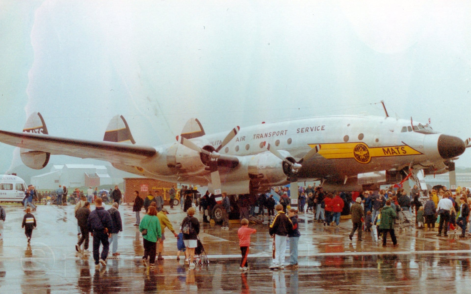 Side view of a 4 propellor-engined, triple-tailed, military transport aircraft in a bare metal finish, parked facing to the right.There is a yellow flash on the nose with a black wireframe globe and black "MATS"  titles under the nose, and black "Military Air Transport Service" titles on the upper fuselage. The tips of the propellors have red, white, and blue stripes, which will make concentric coloured circles when the engines are running. Crowds of people are gathered around and under the plane, some walking around, others standing and looking. The concrete apron they are on is saturated with rain water, puddles reflecting the people and plane. Grey sky fills the top of the frame.