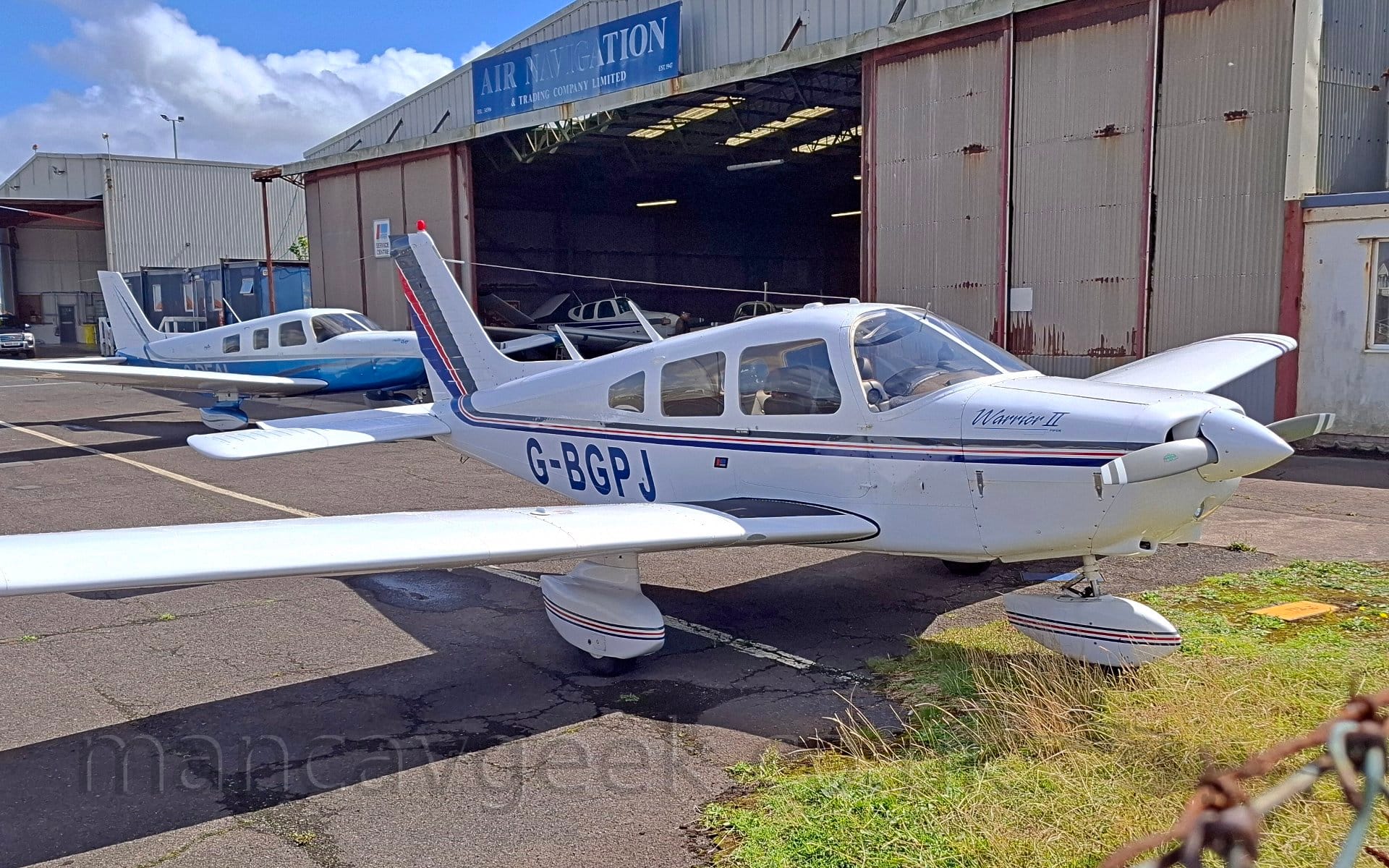 Side view of a white, single propellor-engined light aircraft parked facing to the right. The plane has a grey, red, and blue stripe running along the body under the cockpit windows and up into the tail. The registration "G-BGPJ" is on the lower rear fuselage in blue, while the engine cover on the nose has "Warrior II" titles. There is a large hangar in the background with a selection of light aircraft visible through the open doors, with another parked outside. Blue skies and fluffy white clouds fill the rest of the frame.