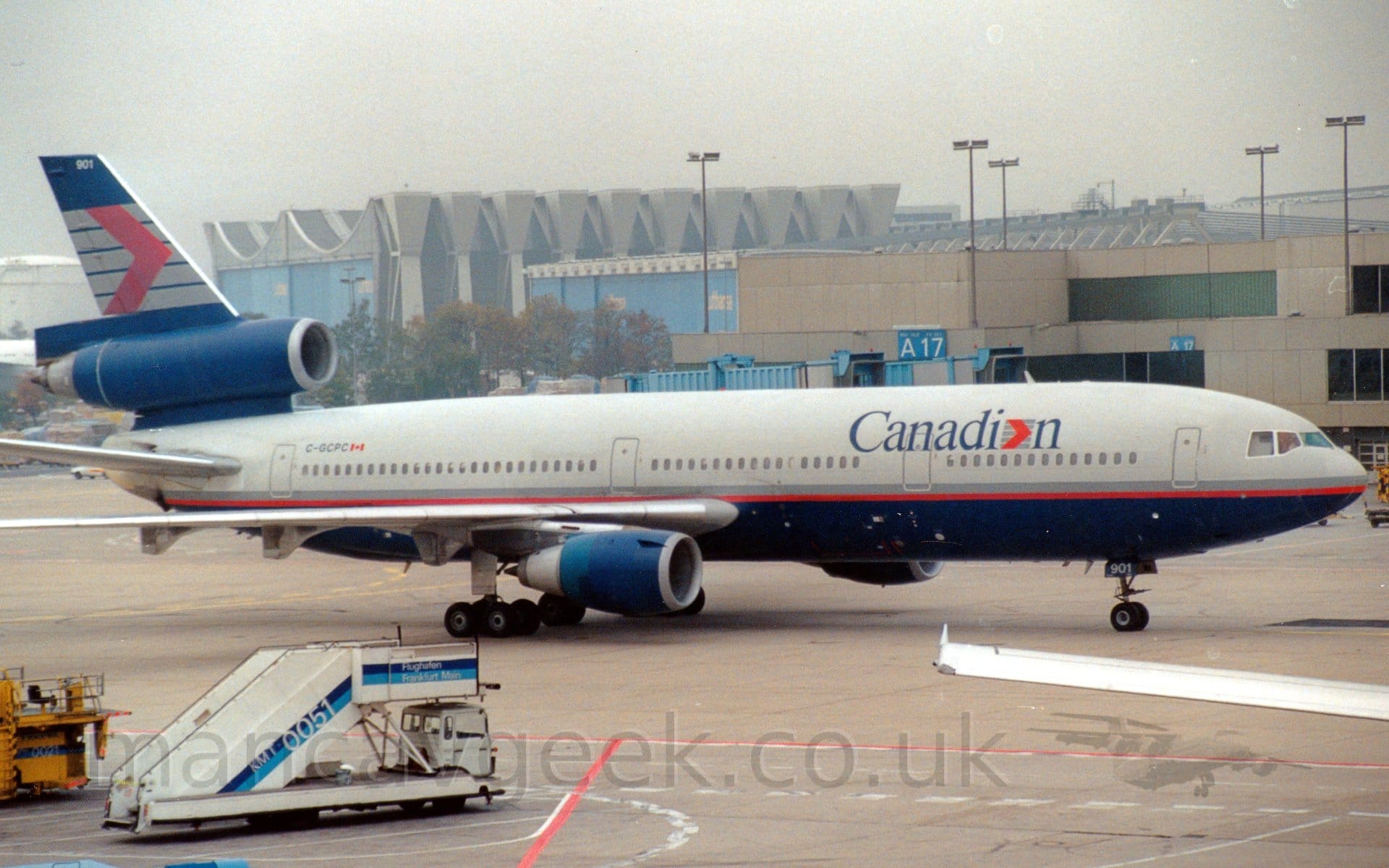 Side view of a grey, 3 engined jet airliner with a dark blue belly and engine pods, and a grey and red stripe running along the body, taxiing from left to right in front of a grey terminal building. The plane has dark blue "Canadian" titles on the upper forward fuselage, the last letter "a" replaces with a red arrowhead overlaid on 5 grey horizontal bars. That same logo appears in a larger form on the dark blue tail. A white set of mobile airstairs is parked on the left of the frame in the foreground, with the wing of another plane on the right. A grey terminal building stretches across the background, with a large grey hanger with pale blue doors is visible on the left.