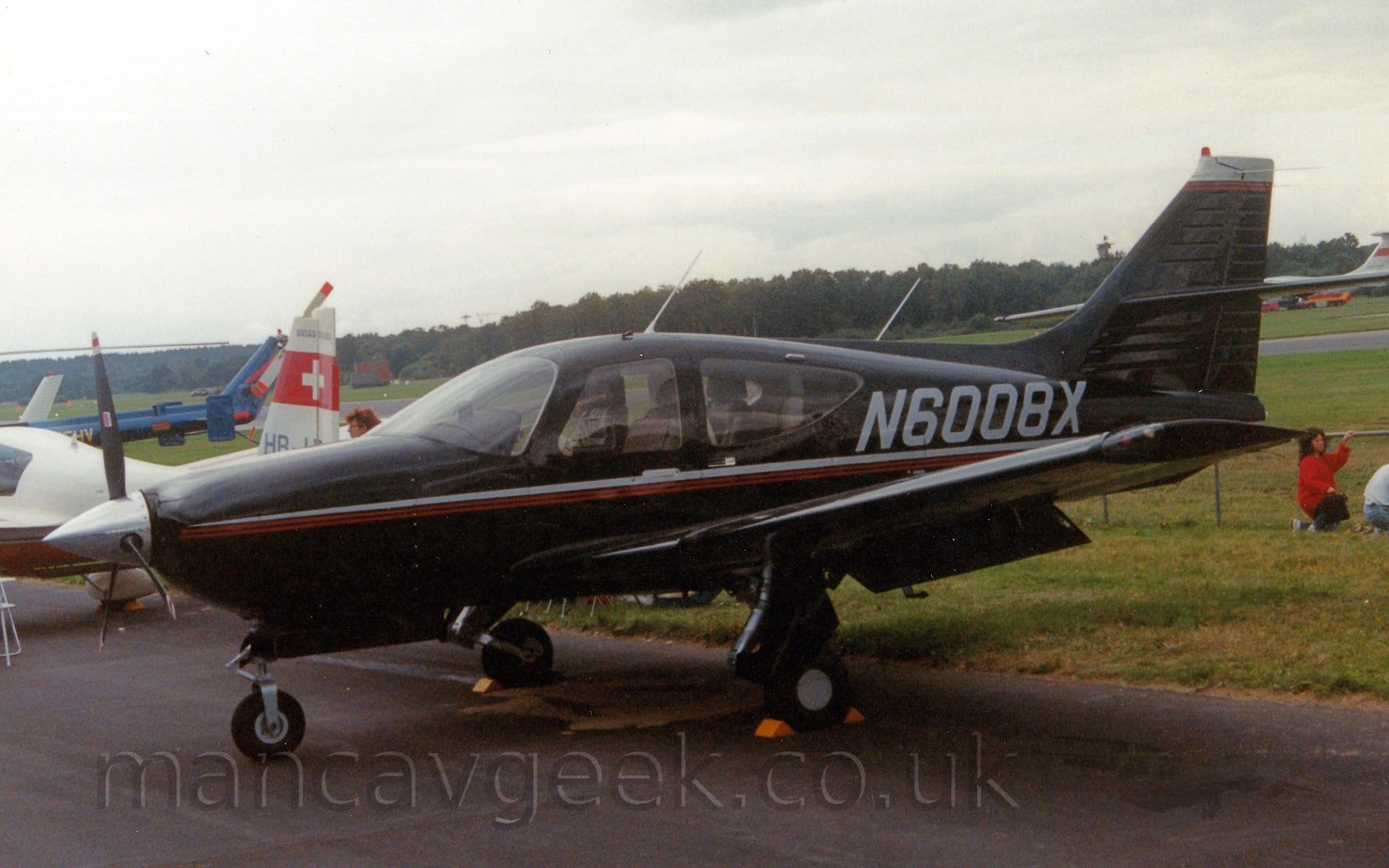 Side view of a black, single engined light aircraft parked on tarmac facing to the left. The plane has a grey and red stripe running along the body from the nose, with the grey registration "N6008X" above the line on the rear fuselage, just aft of the cockpit. The rear fuselage and tail of a white light aircraft with a red stripe containing a white cross on the tail can e seen in the background on the left, with the tail boom of a blue helicopter beyond that. A woman with brown hair and wearing a large red jumper can be seen crouched down next to a metal fence next to a man wearing a white shirt in the background on the right. Grassed areas separated by a black runway lead up to trees in the distance, under grey skies.