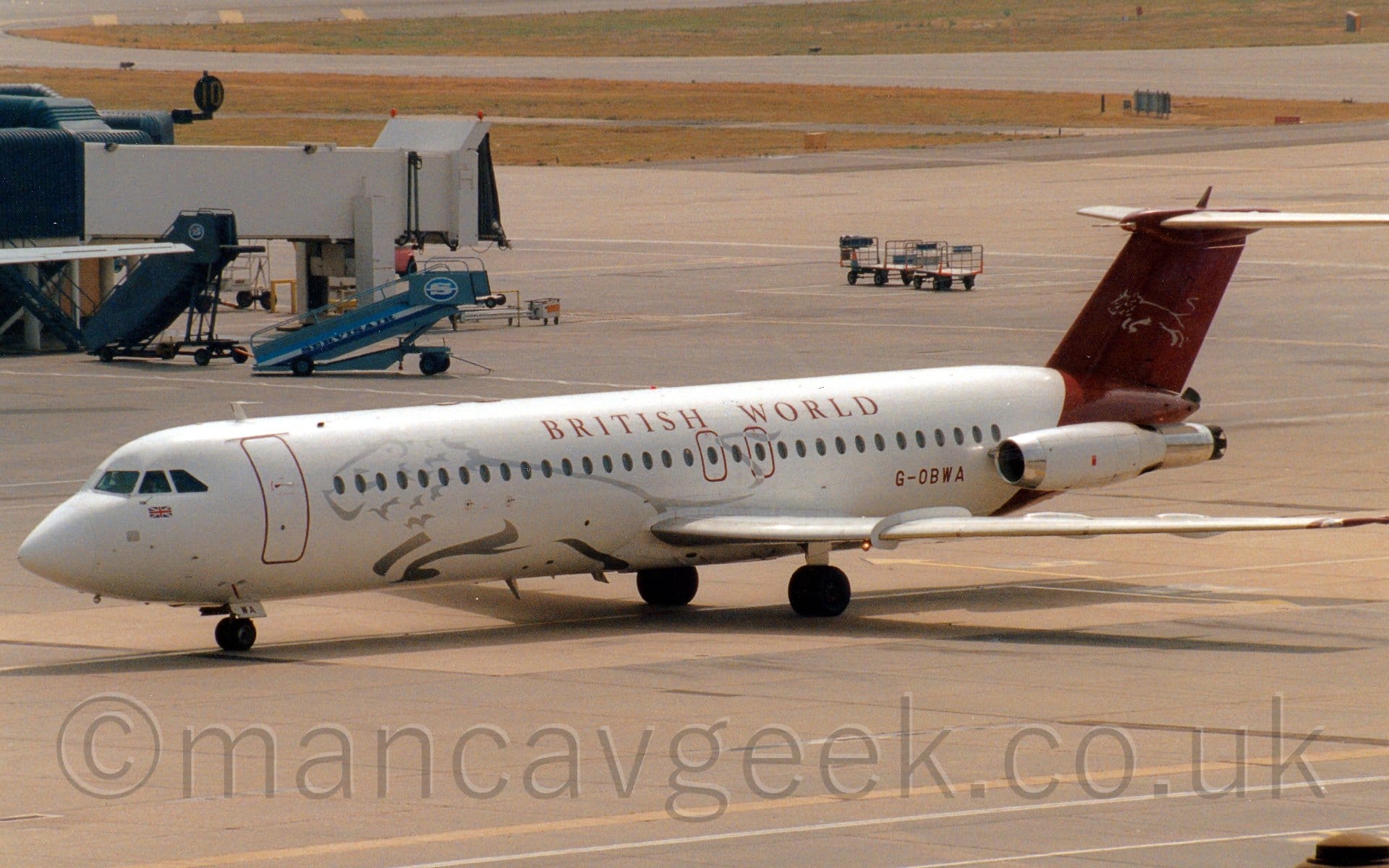 Side view of a white, twin engined jet airliner with the engines mounted on the sides of the rear fuselage, taxiing from right to left. The plane has the silver outline of a leaping lion on the forward fuselage, mauve "British World" titles on the upper fuselage, and a mauve rear fuselage and tail with the silver outline of a leaping lion. Grey apron fills most of the frame, with a white airbridge attached to the edge of a terminal building on the left of the frame. Grassed areas in the distance mark the edges of taxiways at the top of the frame.