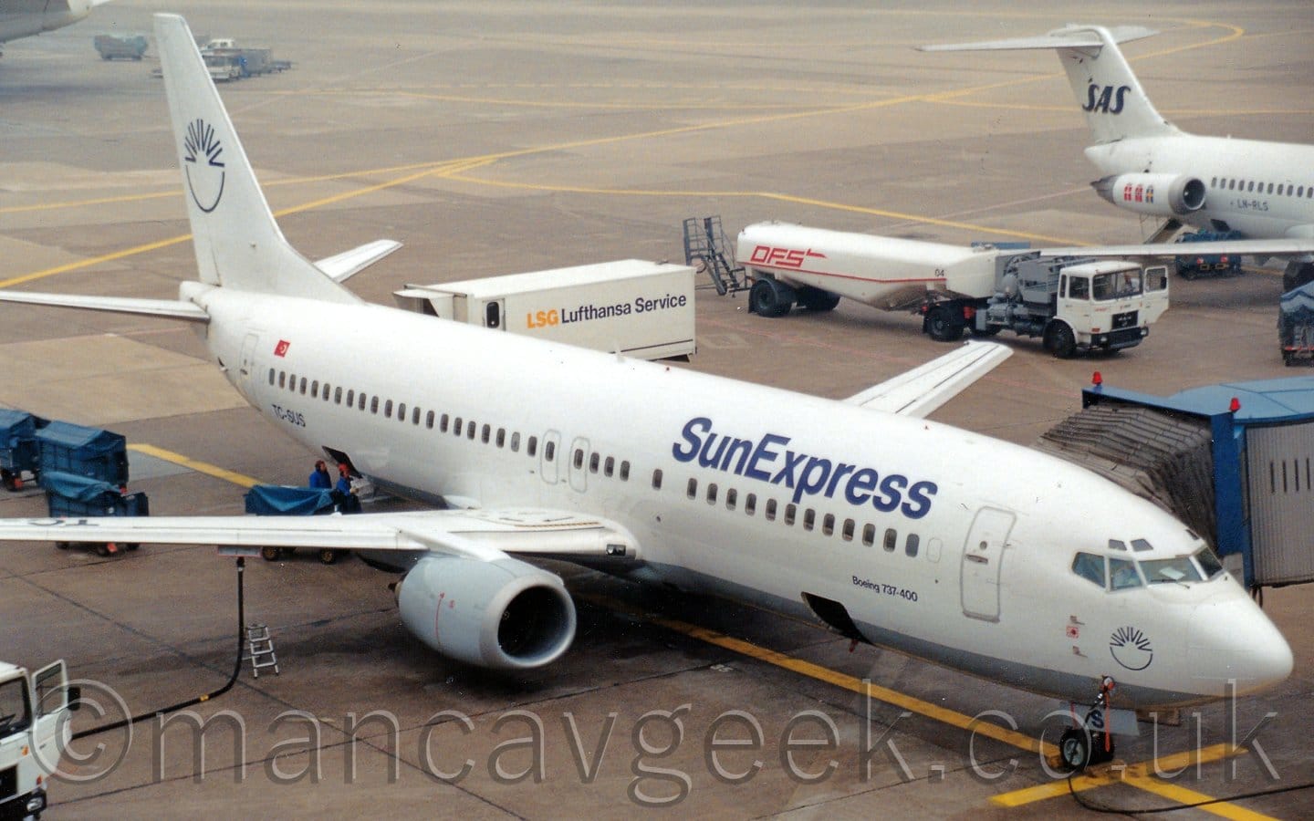 Side view of a white, twin engined jet airliner with a grey belly parked facing to the right by an airport terminal building. A grey airbridge is connected to the planes forward left door. There are dark blue "SunExpress" titles on the upper forward fuselage. There is the blue outline of a sunburst on the tail, a loge repeated on the forward fuselage under the cockpit. Concrete apron fills the rest of the frame in the background, with various trucks, fuel tankers, and baggage carts scattered around, with the rear fuselage of another white jet airliner on the top right.