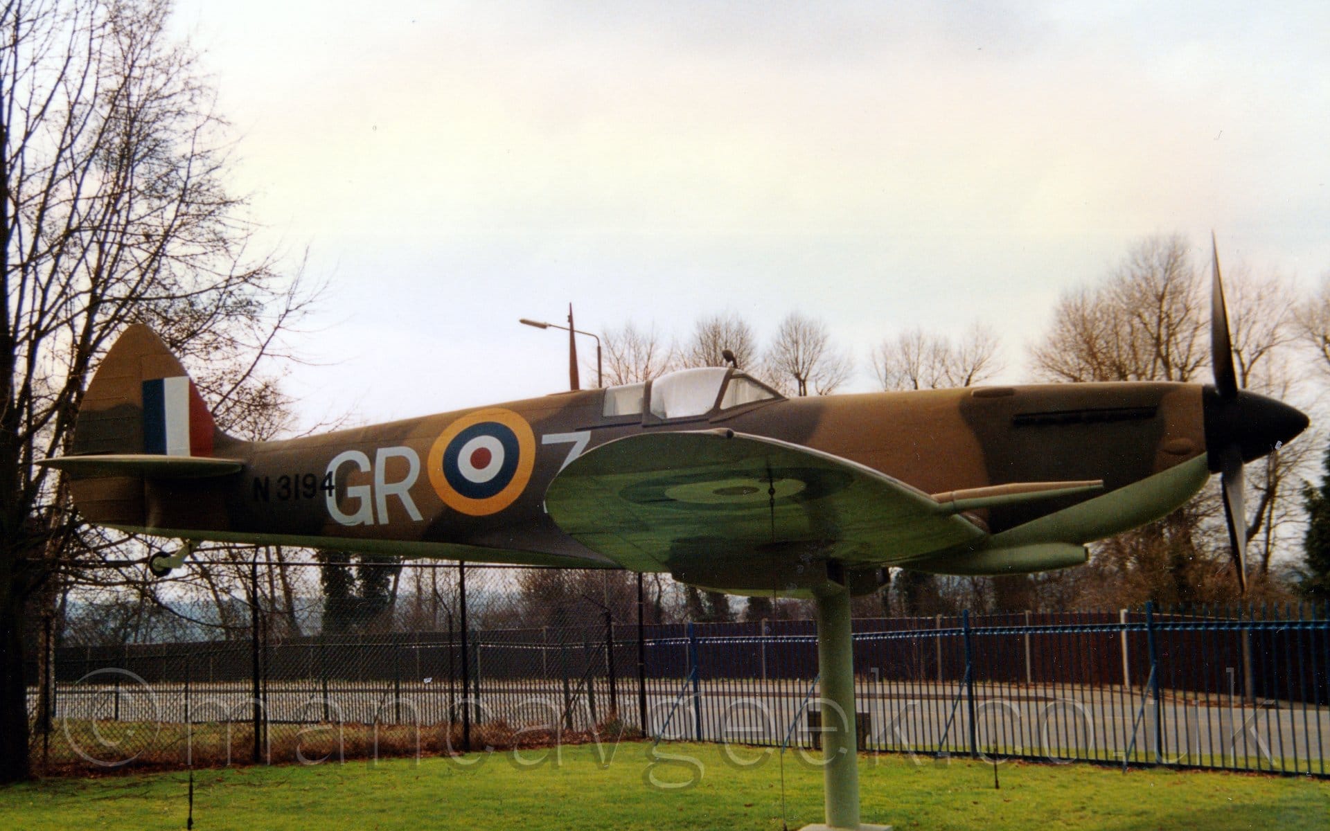 Side view of a replica World War 2-era fighter plane mounted on a pole in a flying position. The plane is covered in a two-tone brown camouflage scheme, except for the belly and undersides of the wings, which are a creamy blue colour. There is a large yellow, blue, white, and red roundel on the rear fuselage, with large white letters "GR" on the left and "Z" on the right. The serial "N3194" is on the rear fuselage in black, the "4" overlaid on the large white "G". A creamy blue pole comes down from the lower fuselage in the middle of the wings, meeting a concrete-filled hole in the ground. Green grass fills most of the foreground under the plane, with black and blue fences separating it from a nearby tree-lined street. Grey skies fill the rest of the frame.