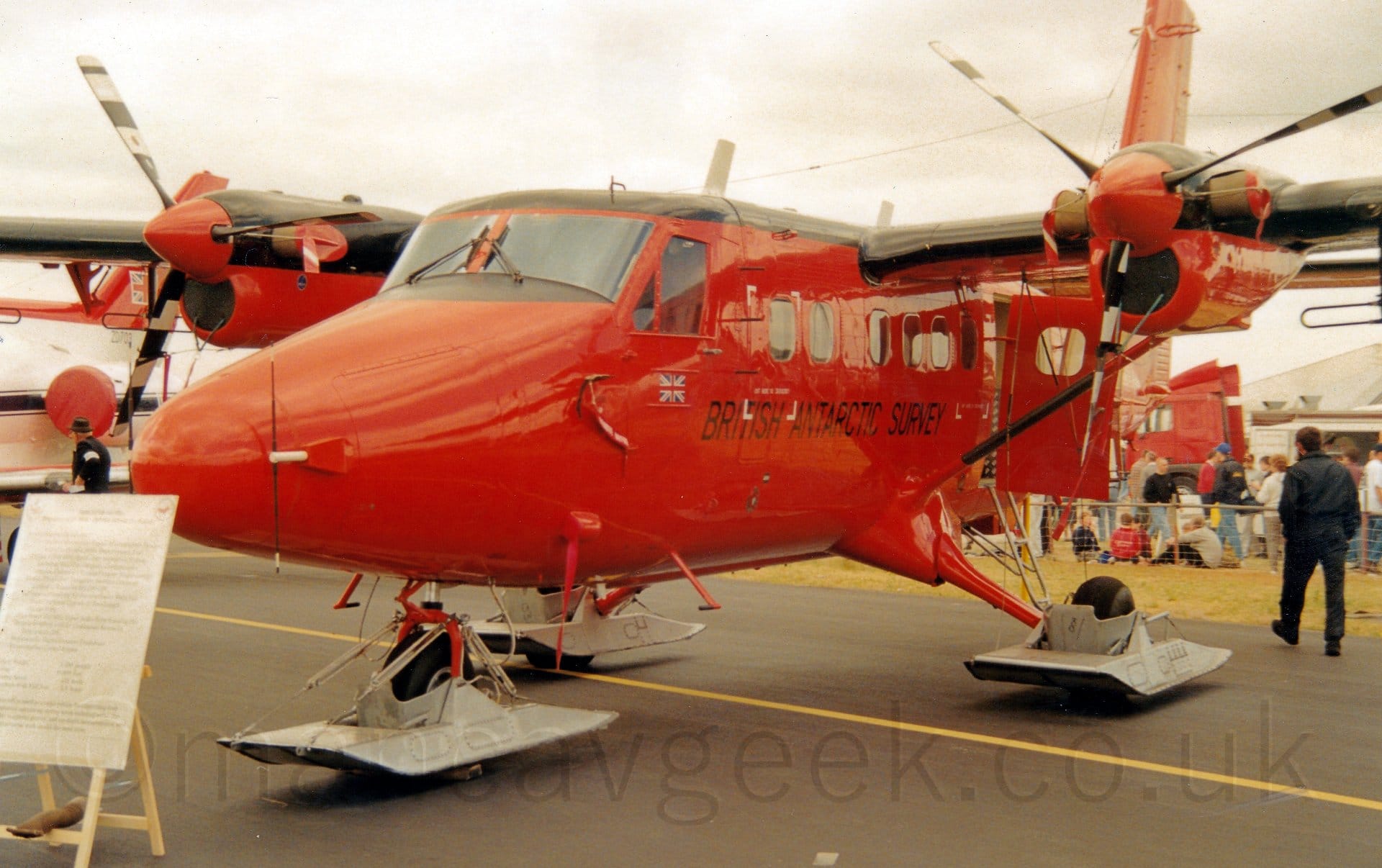 Front side view of a bright red, high-winged, twin propellor-engined parked facing to the left and slightly towards the camera. The plane's roof and the tops of the wings and engines are a shiny black, and black "British Antarctic Survey" are on the lower forward fuselage under the passenger cabin windows. A door in the rear fuselage is hanging open. Metal skids are mounted on the undercarriage, the wheels poking through the bottom. There is a white board on a wooden easel which has indecipherable text about this plane which partially obscures the nose on the left of the frame. Crowds of people behind a low metal fence are visible in the background, under flat white skies.