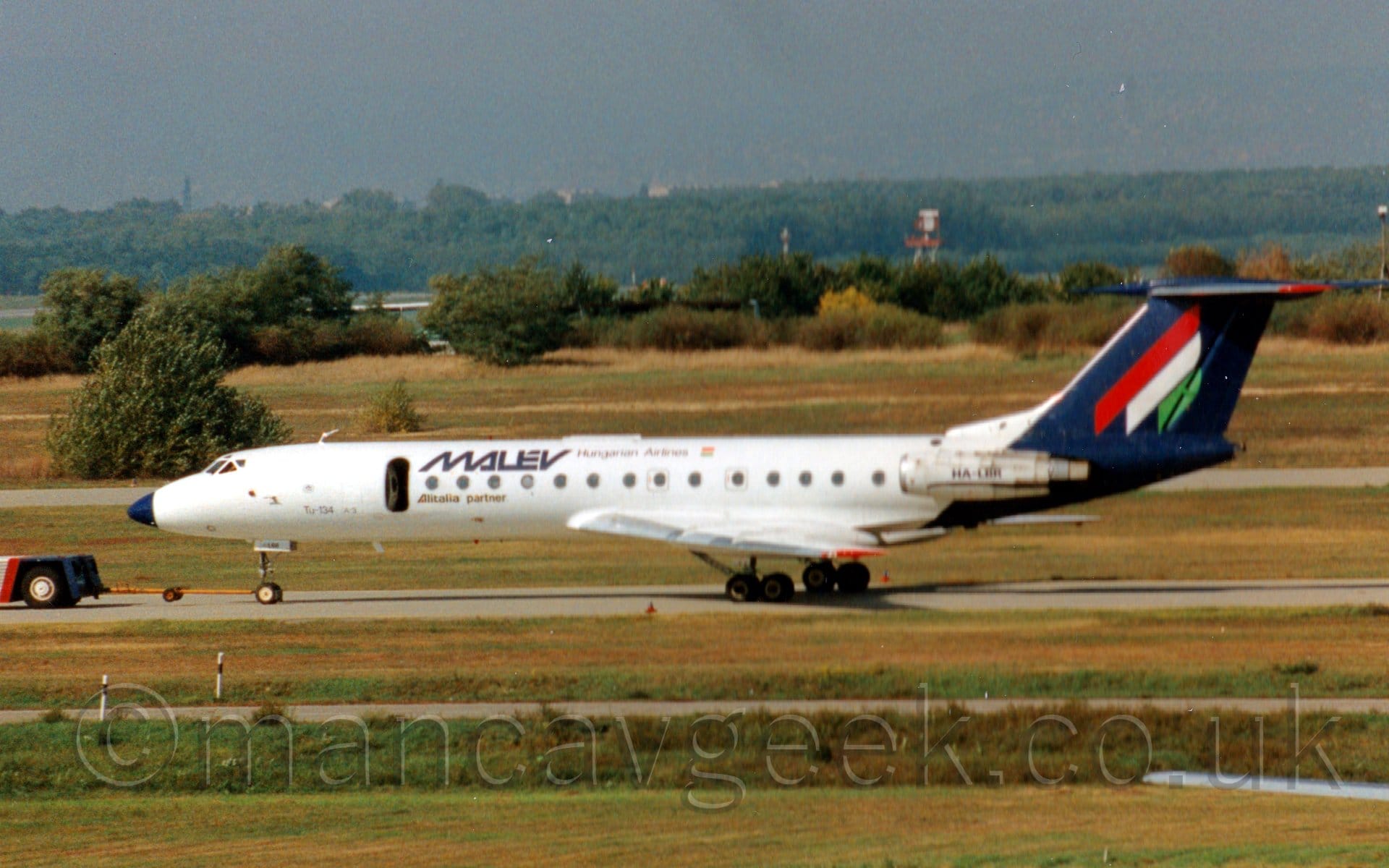 Side view of a white, twin engined jet airliner with the engines mounted on the sides of the rear fuselage, being towed from right to left by a low slung truck vanishing out of frame on the left. The plane has blue "Malev" titles on the upper middle fuselage, just aft of an open door, and a dark blue rear fuselage and tail with red, white, and green diagonal stripes vaguely resembling a letter "M". Yellowing grass fills the foreground, and stretches off into the distance in the background, giving way to trees, under grey-blue skies.