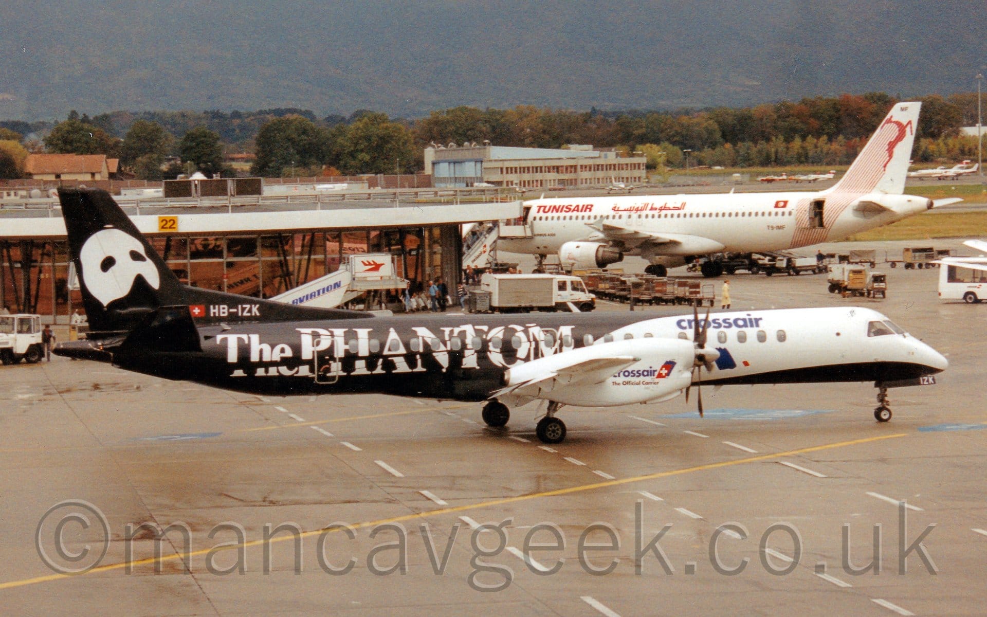 Side view of a white and black, twin propellor-0engined airliner taxiing from left to right through a busy airport scene. The plane has a white forward fuselage with a black belly, and white engine pods on the wings with blue "Crossair" titles in front of the wings. The rear fuselage is black, with large white "The Phantom" titles in a fractured font. The black tail has a large, white mask that would cover the top half of someones face, the lower part of the nose and the mouth being uncovered. The registration "HB-IZK" is on the lower part of the tail in white. Grey apron fills the foreground, with a larger, white and red twin engined jet airliner on the right of the frame parked next to a circular terminal building on the right. White trucks, vans, and mobile airstairs litter the background, along with rows of open baggage carts. A variety of light aircraft and bizjets are parked on the far side of the runway in the distance on the right of the frame, with a large grey concrete hangar in the middle, with trees stretching across the frame beyond that, with a tree covered mountain rising to fill the rest of the frame at the back.