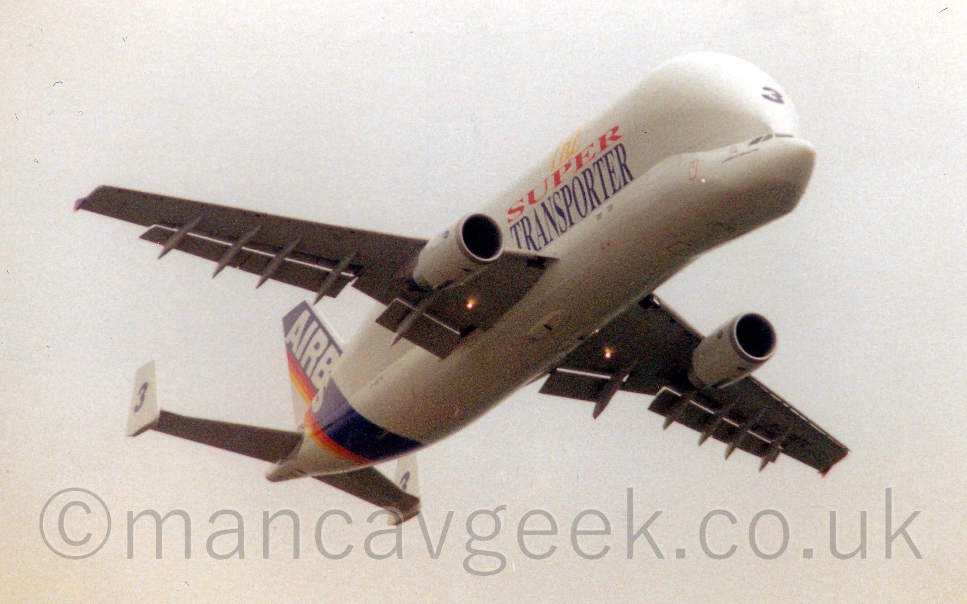 Low side view of a white, twin engined jet airliner flying from left to right at a very low altitude. The undercarriage is retracted, and large flaps are deployed from the rear of the wings. The plane's cockpit is obviously in a different place than normal, being lower than the main fuselage, while the bulbous, extremely wide and tall upper fuselage bulges out significantly wider than the main part of the body at the bottom. Three words adorn the swollen fuselage - "The Super Transporter", in yellow, red, and blue, respectively. A diagonal blue band with a red and orange stripe on the rear edge covers the tail and rear fuselage, with the word "Airbus" overlaid in white. There is a large, blue, number "3"3 facing forwards on the over-sized upper fuselage, above the cockpit. Grey cloudy sky fills the rest of the frame in the background.