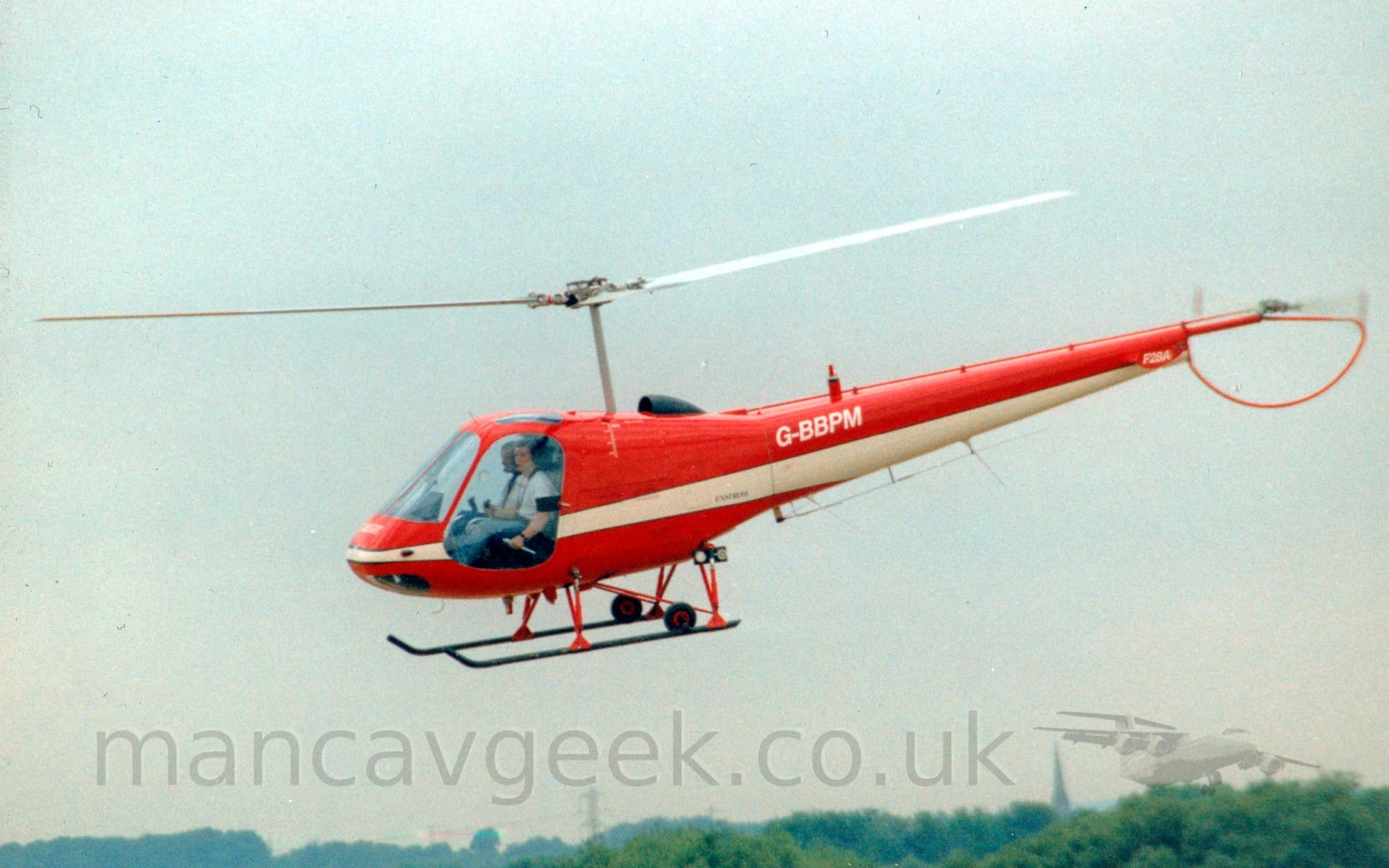 Side view of a red 2 seat helicopter flying from right to left at a low altitude. There is a thick cream stripe running along the body and along the lower side of the tail boom, obviously skipping the glass cockpit doors, and the white registration "G-BBPM" on the upper side of the tail boom. There are 2 people visible in the cockpit through the glazed doors. The tops of trees in the distance are visible at the bottom of the frame, with the rest of the background filled with grey skies.