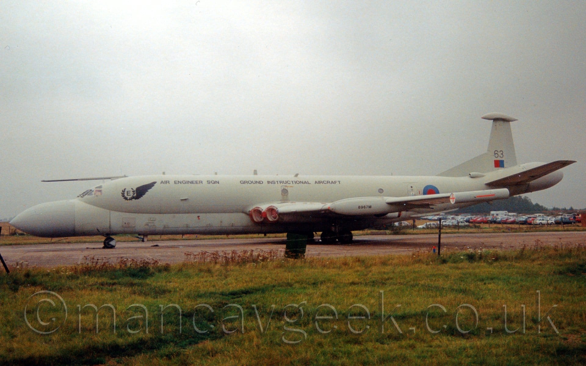 Side view of a grey, 4 engined military jet patrol aircraft parked facing to the left. The plane's upper fuselage is a dark grey, the lower fuselage and the bulbous nose and tail-coner are a lighter grey. The engines are buried in pairs in the wing root, with red covers over the inlets. There are black "Air Engineer Sqn" and "Ground Instructional Airframe" titles on the upper fuselage in black. There is a red and blue roundel on the rear fuselage, and a red and blue fin flash on the tail, under the number "63". Green grass fills the foreground, with cars parked in a field in the background, visible under the planes tail, under grey skies.