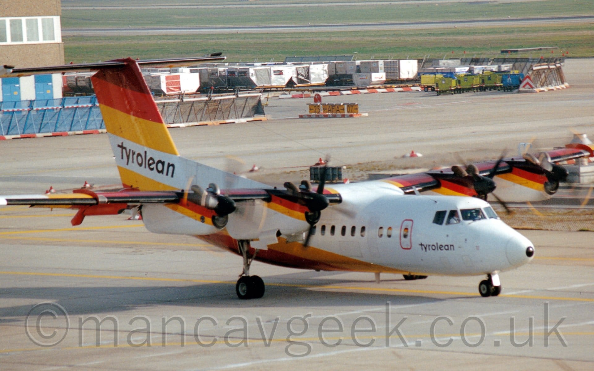 Side view of a white, hi-winged, 4 propellor-engined airliner taxiing from left to right. The plane has thick dark umber, red, and yellow diagonal stripes running along the top of the tail and the front of the top of the engine pods, with the same stripes but in a reversed order running down the base of the tail and rear fuselage, curving under the body in front of the wing. There are large black "Tyrolean" titles on the white part of the tail, repeated in a smaller form under the cockpit windows. Grey taxiway fills the bottom of the frame, with more taxiways bordered by baggage carts and large luggage containers in the background, with acres of green grass leading off to the distance at the top of the frame.