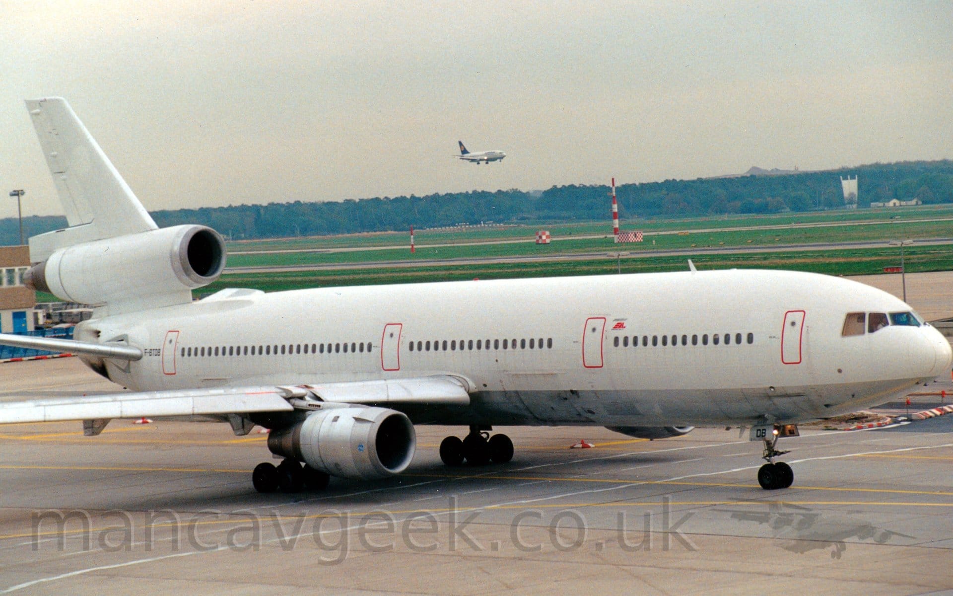 Side view of a white, 3 engined jet airliner with a silver belly taxiing from left to right. The plane is almost entirely blank, only red outlines on the doors, the registration "F-BTDB" on the rear fuselage, and a small red "EAL" logo in front of the mid-forward door providing a break from the "polar bear" livery. In the background, green grass leads off to trees in the distance under a grey sky, with a white, twin-engined jet airliner with a blue tail on final approach to a runway visible in the mid-distance.