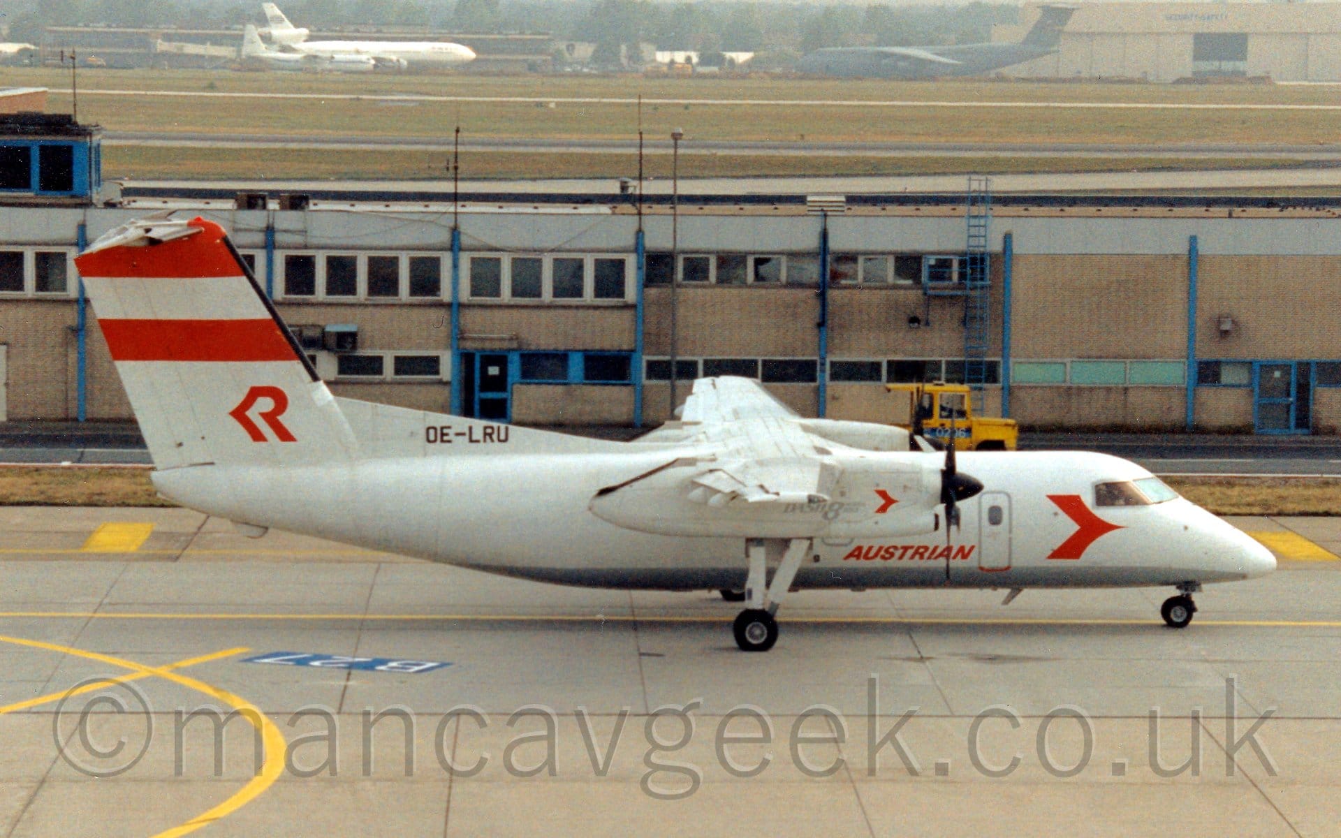 Side view of a white, high-winged, twin propellor-engined airliner taxiing from left to right. The plane has a stylised red arrowhead on the forward fuselage, just below and aft of the cockpit, and red "Austrian" titles on the lower fuselage. The same arrowhead is on the white engine pods, in a slightly smaller form. There are 2 red stripes at the top of the tail, and a red stylised upper-case letter "R" at the base, with the registration "OE-LRU"  at the bottom in black. Grey taxiway fills the foreground, with a 2-storey sandy brown and grey building stretching across the frame in the background. In the distance, acres of grass lead up to cargo planes in front of hangars  at the top of the frame.