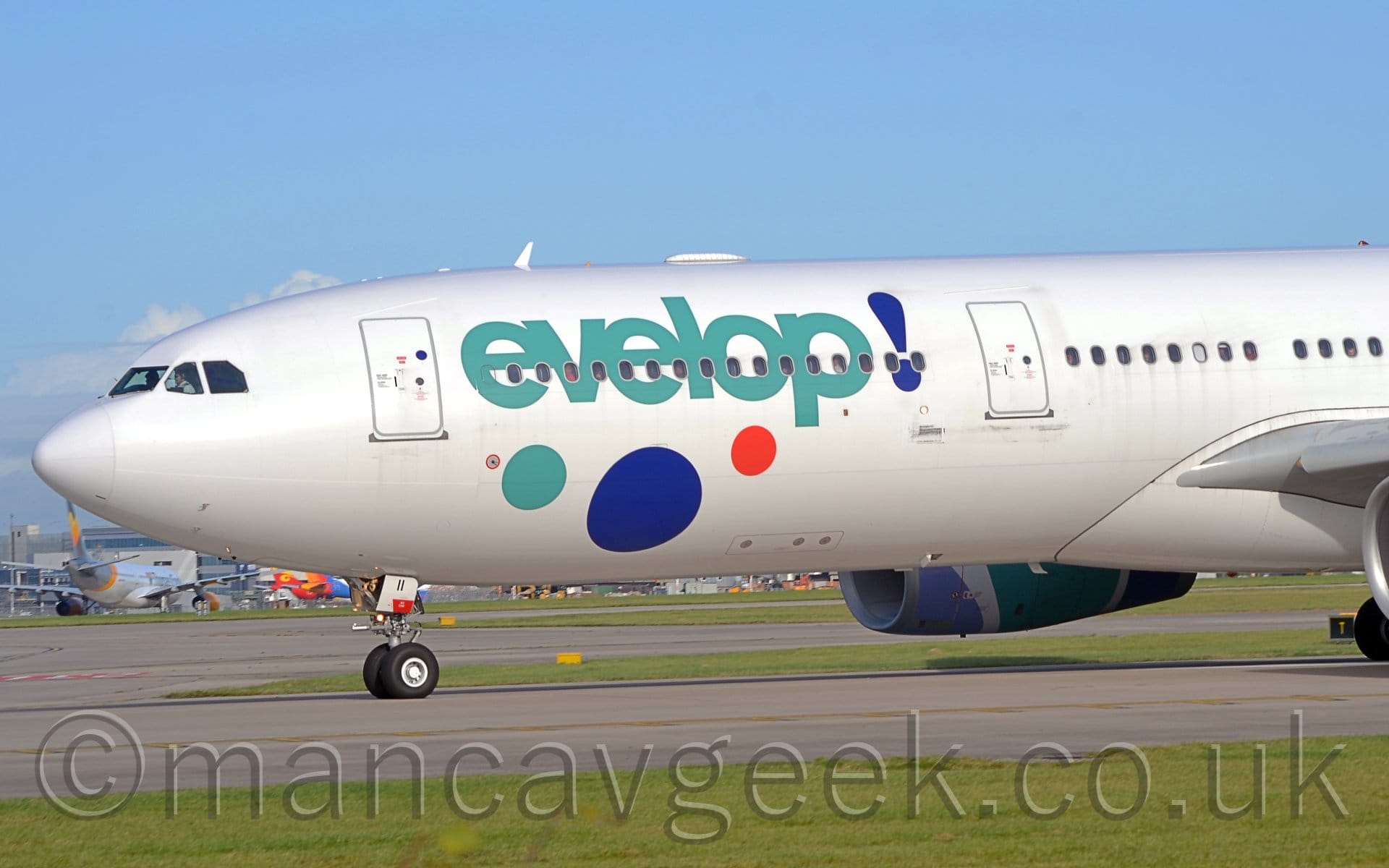 Close-up of the forward fuselage and nose of a white, twin engined jet airliner taxiing from right to left. There are large, aqua-marine "Evelop" titles on the forward fuselage, followed by a large purple exclamation mark, and with 3 large dots (aqua-marine, purple, orange) underneath. The engine mounted on the wing on the other side of the plane is partially visible under the fuselage, it's cowling painted pastel purple, aqua-marine, and dark purple, with white stripes. Green grass fills the foreground with more grass visible under the plane leading up to a terminal building in the distance. Blue skies fill the rest of the frame.