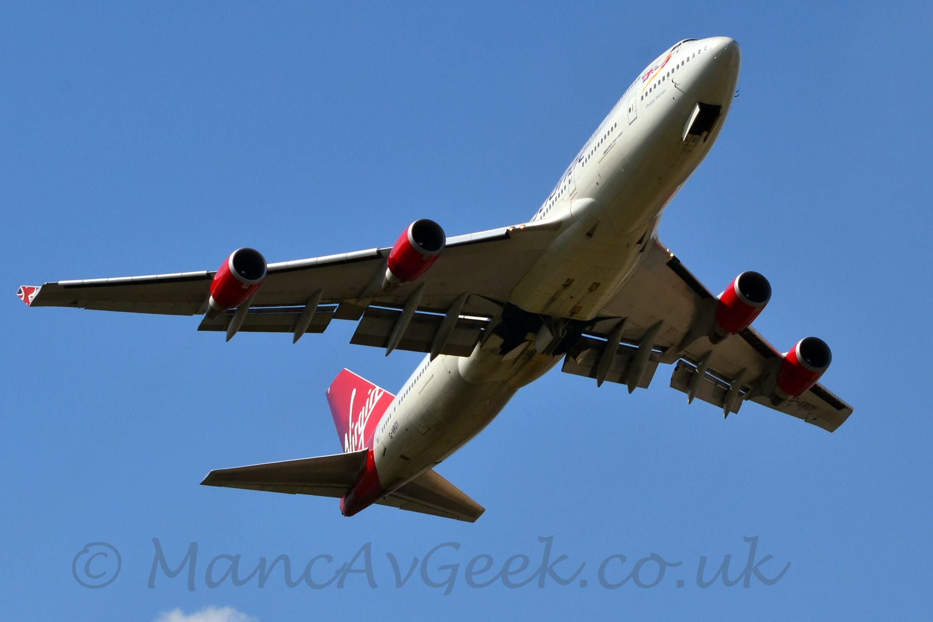 View from below of a very large, white, 4 engined jet airliner flying from left to right at a very low altitude, with its undercarriage almost fully retracted, flaps deployed from the rear of the wings, and the nose raised sharply, all suggesting it has just taken off. The tail and the 4 engine pods slung under the wings are a bold, bright scarlet red, with white, seemingly handwritten "Virgin" titles on the tail. The registration "G-VROY" is on the lower rear fuselage and under the end of the left wing. Blue sky fills the rest of the frame.