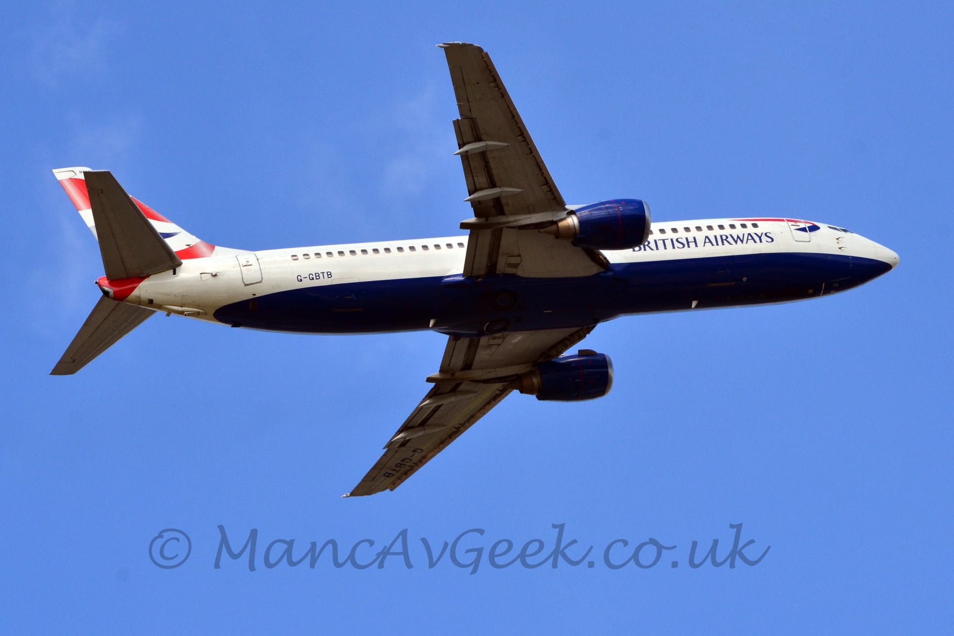 Low side view of a blue, twin engined jet airliner with a deep blue belly flying from left to right at a very low angle, with undercarriage securely tucked away, flaps extended from the rear of the wings, and with the nose raised, suggesting it has just taken off. The plane has blue engine pods, blue "British Airways" titles on the lower forward fuselage, and a blue and red twisted ribbon un the upper forward fuselage, just aft of the cockpit. There are red and blue wavy stripes on the tail. Blue sky fills the rest of the frame.