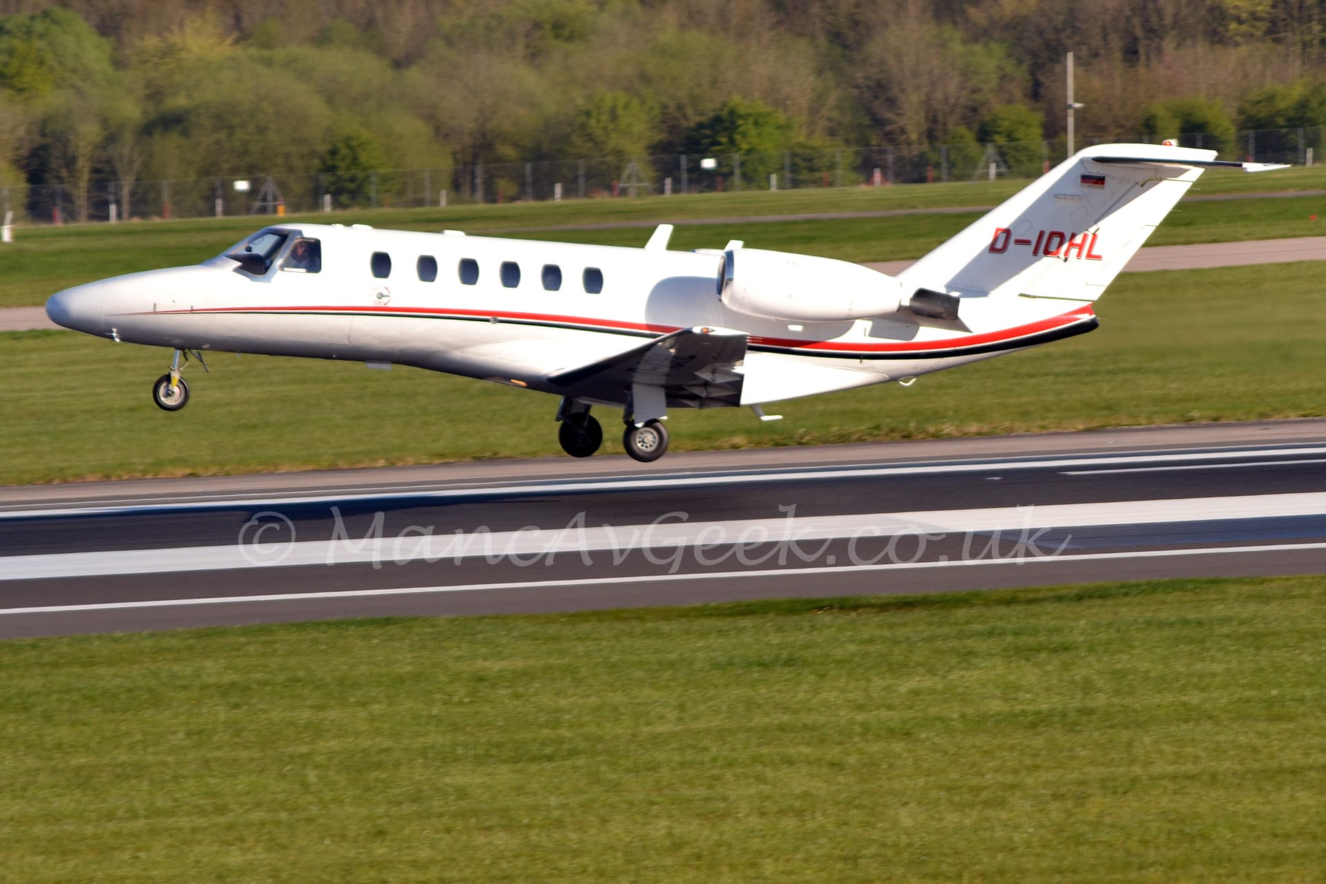 Side view of a white, twin engined biz-jet with the engines mounted on the rear fuselage, flying from right to left at a very low altitude, it's main wheels just a couple of metres above the black runway. The plane has flaps slightly extended behind the wings, and the nose raised significantly, suggesting it has just taken off. There is a thin red and black stripe running along the body, and the registration "D-IOHL" on the tail in red with a black drop shadow. Green grass fills the foreground, with more grass in the background leading up to trees stretching across the frame in the distance.