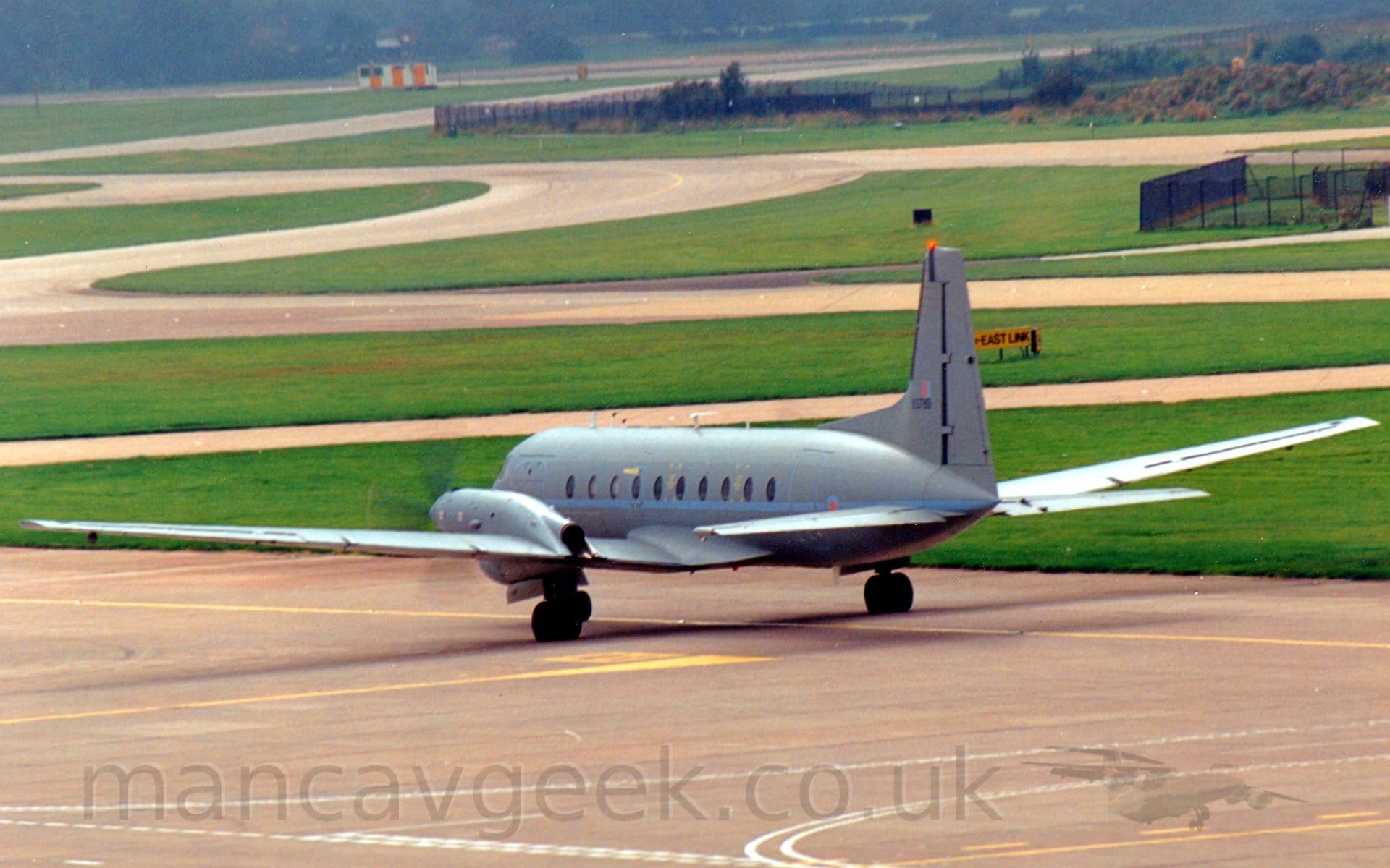 Rear side view of a grey, twin propellor engined military transport aircraft taxiing away from the camera and off to the left. A pale blue stripe runs along the fuselage, just under the passenger cabin windows, with a pale blue and red roundel on the rear fuselage. The serial "XS789" is on the tail in black, below a small, pale red and blue fin flash. Concrete apron fills the foreground, with green grass marking the edges of multiple taxiways winding their way through the background, with trees in the distance.