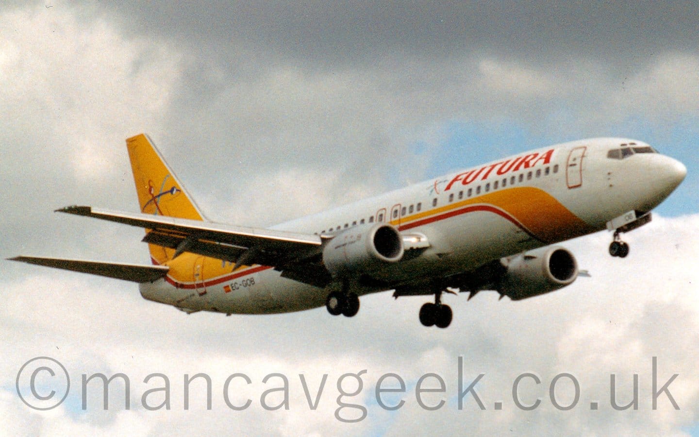 Slightly grainy side view of a white, twin engined jet airliner flying from left to right at a very low altitude, with it's undercarriage lowered, and flaps deployed from the rear of the wing, suggesting it is just about to land. There is a yellow and red stripe coming from under the belly, along the fuselage, and up to fill the tail, which has a logo which resembles 3 electrons (red, dark blue, light blue) in different orbital inclinations spinning around a common centre. There are red "Futura" titles on the upper forward fuselage, just ahead of a smaller version of this same logo. Leaden grey skies with slashes of blue fill the rest of the frame.