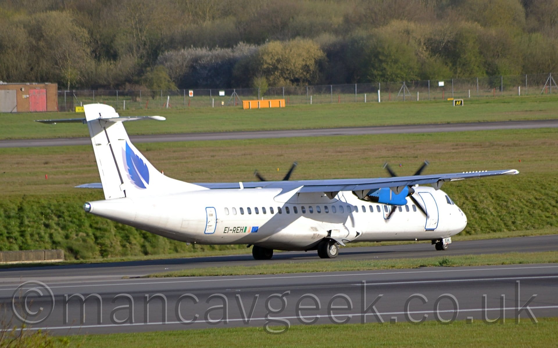 Side view of a white, high-winged, twin propellor-engined airliner taxiing from left to right on a black taxiway. The plane has a blue leaf with a grey drop-shadow on the tail, the registration "EI-DEH" on the lower rear fuselage next to Irish and EU flags, and blue engine pods under the wings. A strip of green grass stretches across the bottom of the frame, with another strip of grass separating the runway and taxiway. More green grass in the background lead up to trees stretching across the frame in the background.