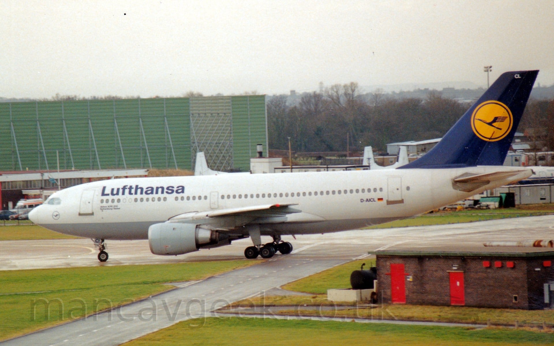 Side view of a white, twin engined jet airliner taxiing from right to left. The plane has a grey belly, dark blue "Lufthansa" titles on the upper forward fuselage, and a dark blue tail with a yellow circle in the middle containing a stylised blue flying bird. The registration "D-AICL" is on the lower rear fuselage in black, next to a small German flag. A low, dark brick building is in the foreground on the right, almost entirely surrounded by grass. In the background, the tails of 3 white planes can be seen over the top of this plane, scattered amidst several small prefabricated buildings, on the right of the frame, with a large, green metal structure can be seen on the left. Trees in the distance run off to the horizon, where they meet a dismal grey sky.