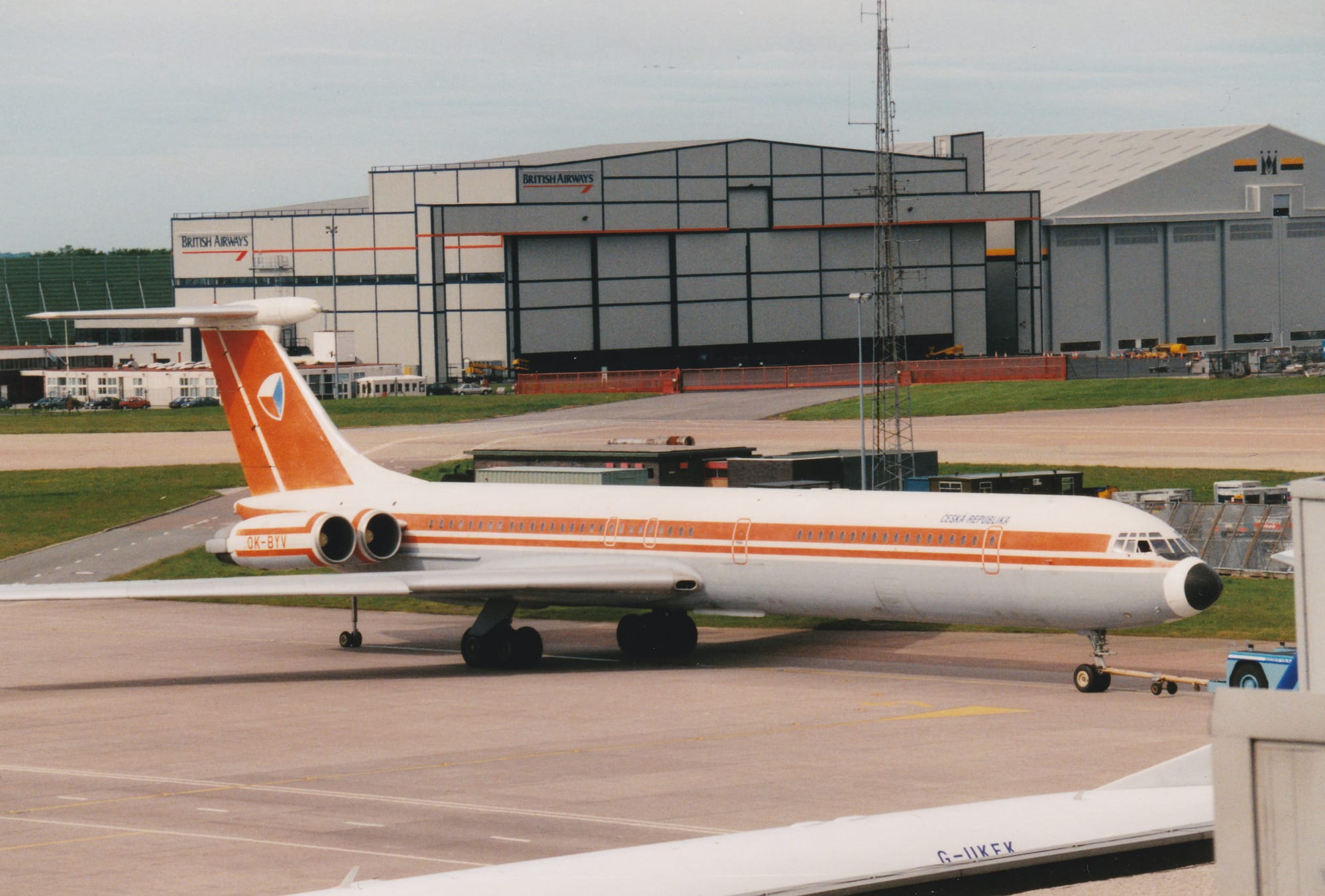 Side view of a white, 4 engined jet airliner with the engines mounted on the sides of the rear fuselage, facing to the right but being pushed back to the left by a low-slung blue truck almost hidden behind a grey building on the left of the frame. There are faded red stripes running along the body, with red stripes on the white engine pods above and below the red registration "OK-BYV", and a red tail with rounded off cube with white, red, and blue faces. There are small, black "Ceska Republica" on the upper forward fuselage. A black pole with wheels at the bottom supports the rear fuselage. Grey apron fills the bottom of the screen, with the top of a white jet airliner and part of a building in the bottom right corner. Strips of green grass mark the edges of a taxiway in the background, with a couple of large grey hangars in the distance, under cloudy grey skies.