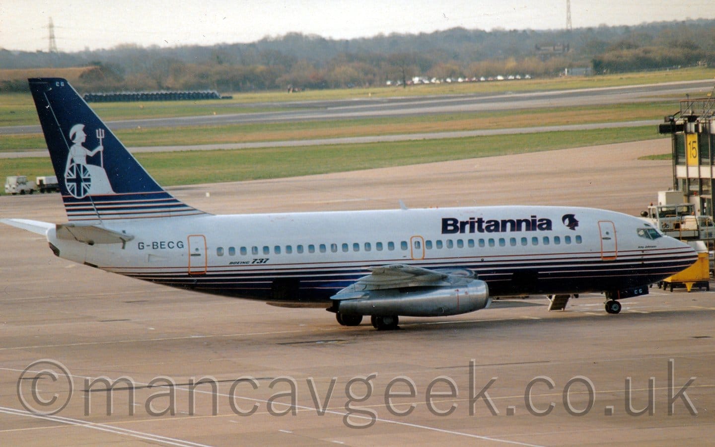 Side view of a white, twin engined jet airliner parked facing to the right. The plane has a series of dark blue stripes on the lower fuselage, getting thicker the lower they go, until eventually the belly is just solid blue. The same happens on the tail, only they merge upwards. There are blue "Britannia" titles on the upper forward fuselage, next to the blue head of a woman wearing an extravagant, plumed helmet. A full, white image of that same woman appears on the tail, wearing white robes, and seated facing to the right, holding a circular shield emblazoned with the Union Flag in her right hand, and a long trident in her left. The position and design of the circular shield actually makes it look as if she is sitting in a wheelchair. The registration "G-BECG" is on the rear fuselage under the tail. Concrete apron fills the foreground, with taxiways and a runway separated by large strips of grass, leading up to trees in the distance under bright but hazy skies.