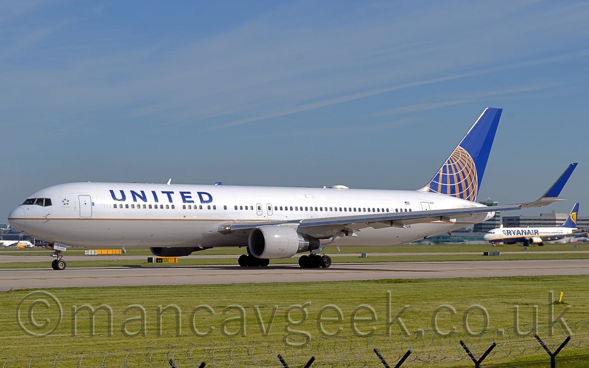 Side view of a white, twin engined jet airliner taxiing from right to left along a runway. The plane has a grey belly and a thin golden stripe running along the body, and a deep blue tail with a white and gold wire-frame globe. There are blue "united" titles on the upper forward fuselage, and blue upturned wingtips. Green grass fills the foreground, with more grass in the background under the plane leading to a large terminal building in the distance. A white and blue jet airliner with "Ryanair" titles can be seen in the distance on the right of the frame. Grey-blue skies fill the rest of the frame.