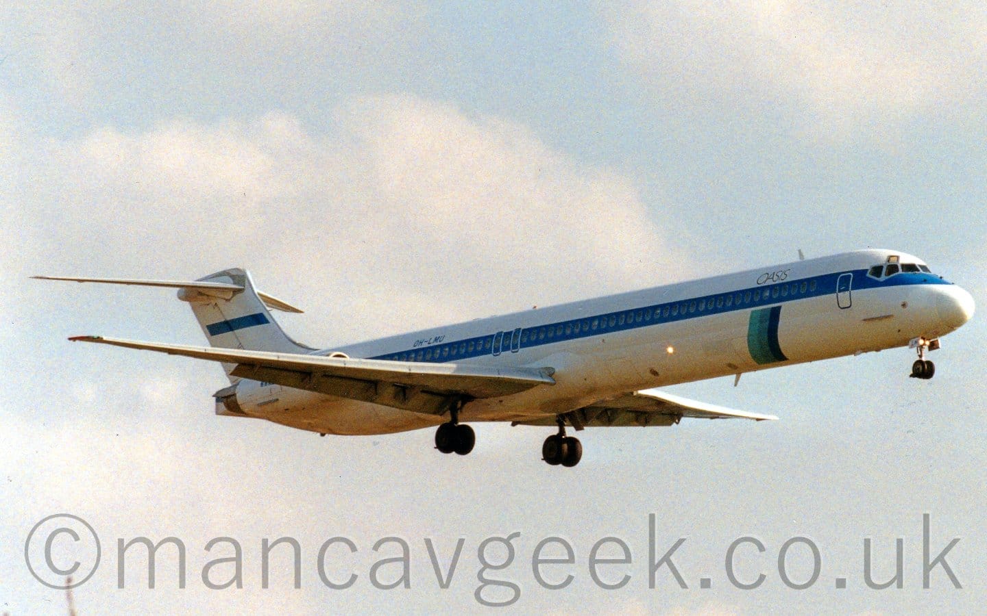 Side view of a white, twin engined jet airliner with the engines mounted on the sides of the rear fuselage, flying from left to right at a very low altitude, with it's undercarriage lowered and flaps extended from behind the wings, suggesting it is about to land. The plane has a thick blue stripe running along the body, covering the passenger cabin windows, and small black "Oasis" titles on the upper forward fuselage. Under the "Oasis" titles, a vertical bar in 3 shades of blue extends down towards the plane's belly. Pale blue sky with hazy white cloud fill the rest of the frame.