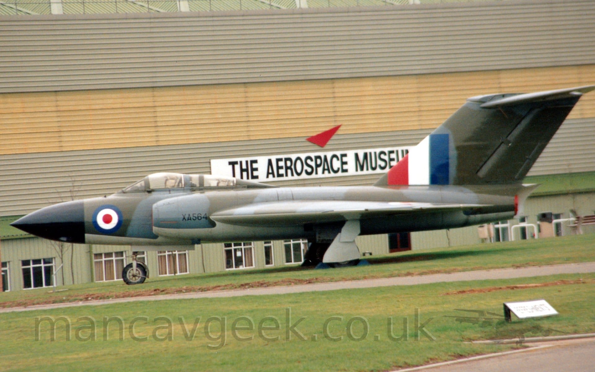 Side view of a twin engined, military fighter jet aircraft parked on grass facing to the left. The plane is mostly covered in a grey-blue and olive green camouflage scheme, with a pale blue belly. There is a large blue, white, and red roundel on the nose, just aft of the black radome, the serial "XA564"in black on the engine inlet under the two cockpit canopies, and a large red, white, and blue fin flash on the front of the tail. Green grass fills the foreground, with a large building in shades of light green and light brown stretching across the frame, a large white banner with "The Aerospace Museum" titles on the side of the building in the middle of the frame.