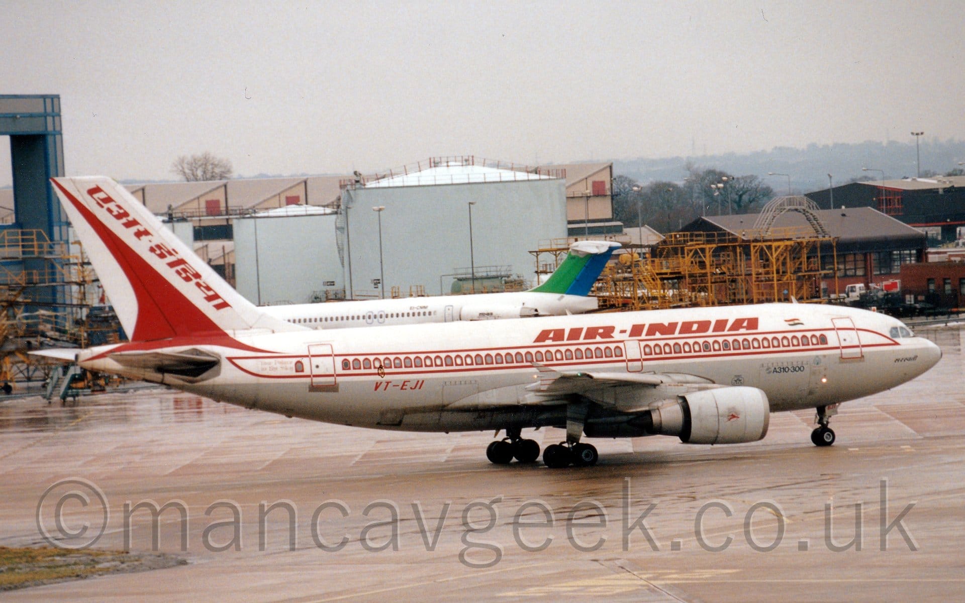 Side view of a white, twin engined jet airliner taxiing from left to right. The plane has a grey belly, and red "Air India" titles along the top of the fuselage. There is the red outline of a stripe running along the body, framing the passenger cabin windows, which are also painted with red arches, to look like windows in a castle or palace. The tail has the red image of a stylised flying bird, along with red titles in an Indian script that translate to "Air India". Grey concrete apron fills the foreground, with a pair of blue fuel storage tanks in the middle of the background, surrounded by large yellow metal frames and black buildings on the right, and a large grey hangar door on the left, under dreary grey skies.