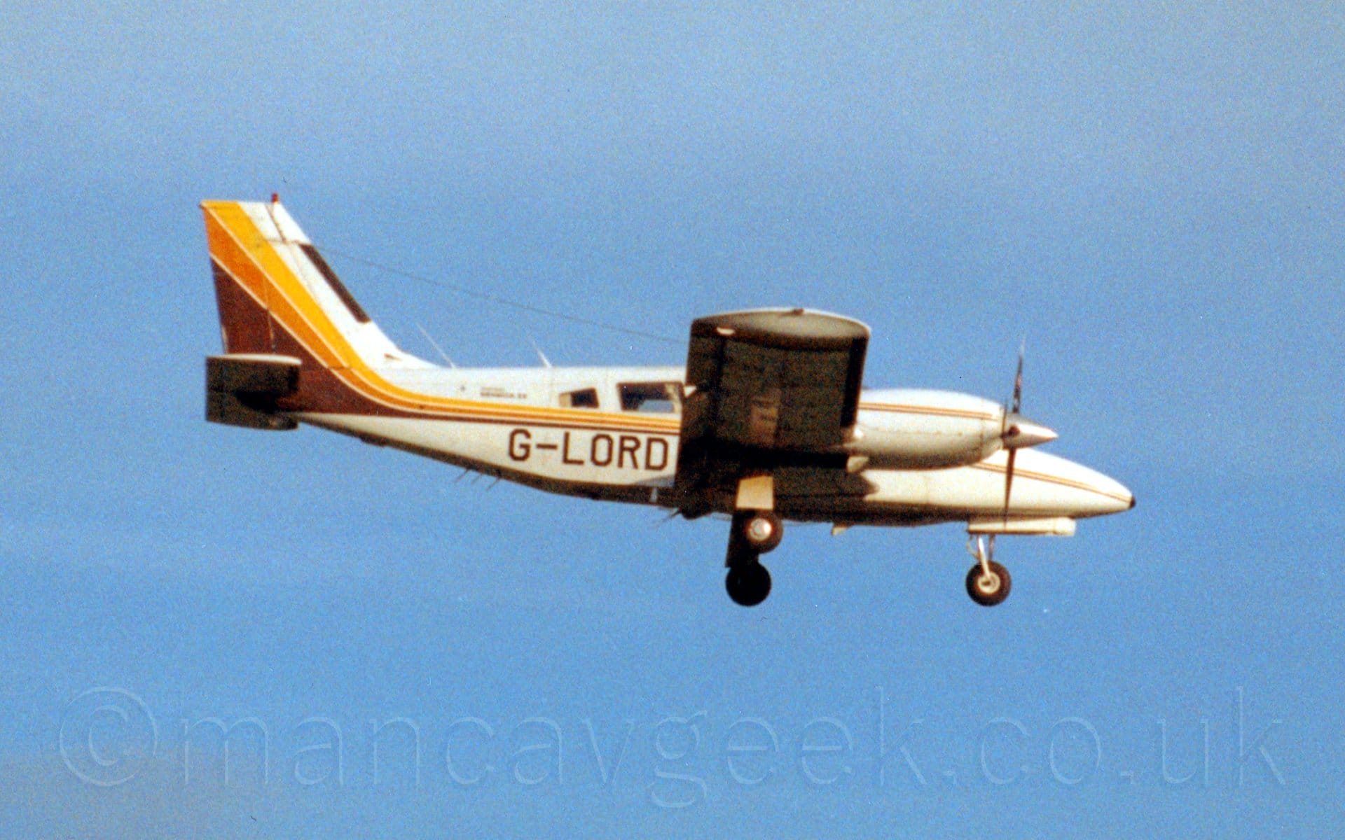 Slightly grainy side view of a cream coloured, twin propellor-engined light aircraft flying from left to right at a low altitude, with the landing gear extended, flaps deployed from the rear of the wing, and the nose down slightly, suggesting it is about to land. There is a thin yellow, orange, and brown stripe running along the body from the nose, sweeping up into the tail as it gets wider towards the rear, with a similar stripe running along the engine cowlings. The registration "G-0LORD" is on the lower rear fuselage in brown. Slightly hazy blue sky fills the rest of the frame.