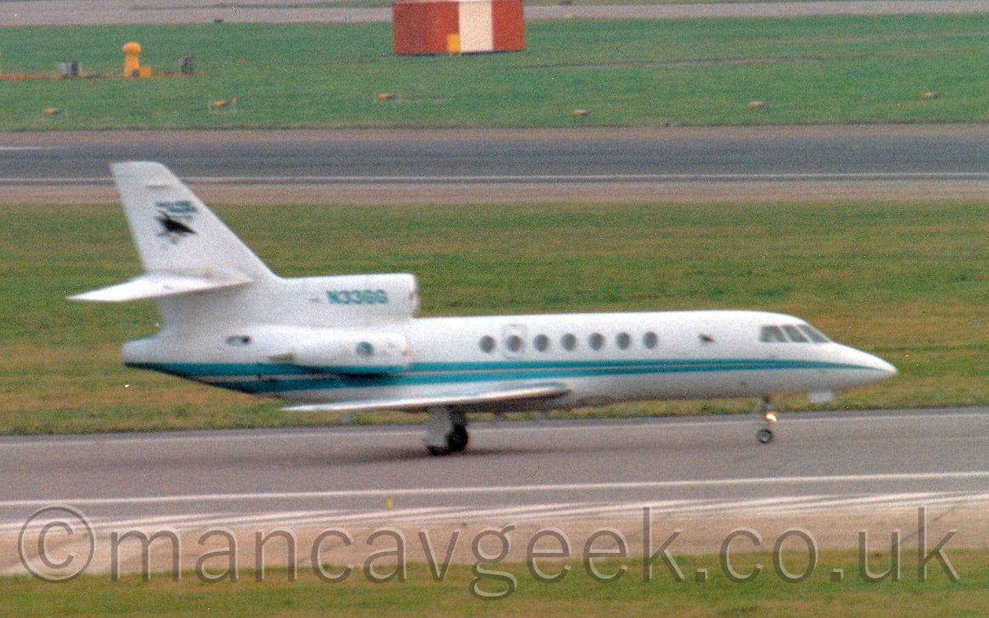 Slightly grainy and fuzzy shot of a long, thin, 3 engined BizJet with the engines mounted on the sides and top of the rear fuselage, taxiing from left to right. There is a pair of thin green stripes running backwards from the nose, getting wider as it moves to the rear. The registration "N33GG" is on the centre engine inlet in the same green colour. A strip of green grass in the foreground marks the edge of the taxiway the plane is moving on, with more grass between the other edge and a runway in the background, with more grass running to the edge of the frame beyond that.