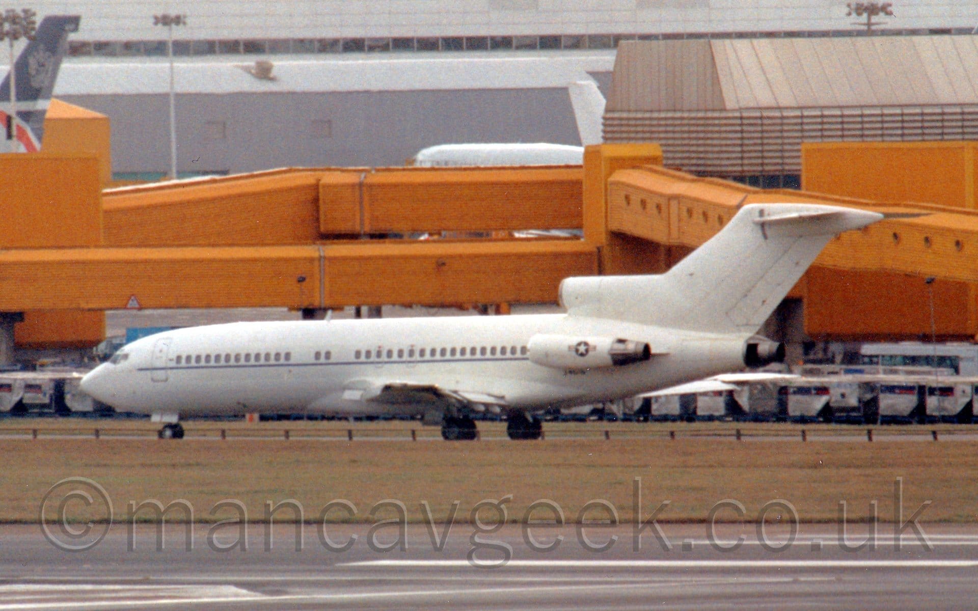 Side view of a white, 3 engined jet airliner taxiing from right to left. There is a thin blue line running along the body under the passenger cabin windows, and a white, 5 pointed star in a blue circle with thin bars on the side on the engine pods mounted on the sides of the rear fuselage. A black runway stretches across the bottom of the frame, with slightly brown grass separating the runway from the taxiway this plane is on. In the background, a large grey terminal building stretches across the frame, with long yellow airbridges stretching in all directions.