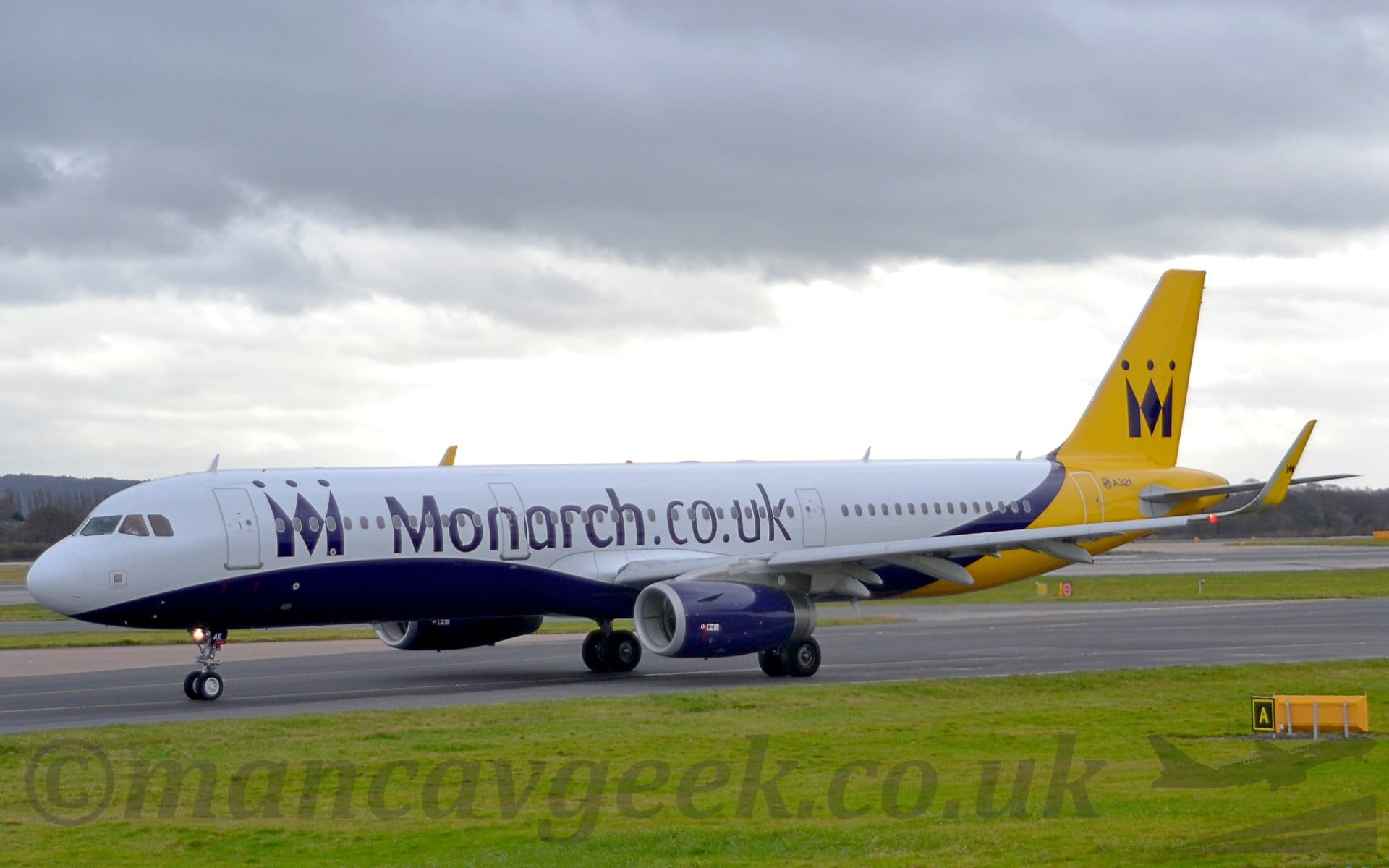 Side view of a white, twin engined jet airliner taxiing from right to left. The plane has a purple belly and engine pods, and a bright yellow rear fuselage and tail with a stylised crown that resembles an upper-case letter "M". A smaller version of that logo appears on the yellow up-turned wingtips, as well as on the forward fuselage, where it is followed by large purple "Monarch" titles. Lush green grass fills the foreground at the bottom of the frame, with more grassed areas marking the edges of taxiways and a runway in the background, leading up to trees on the skyline, under layers of grey cloudy sky.