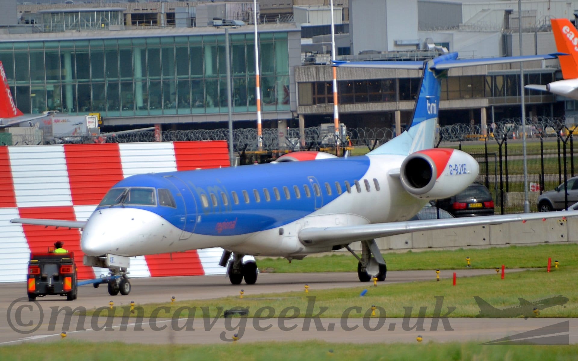 Side view of a white and blue, twin engined jet airliner with it's engines mounted on the sides of the rear fuselage, facing to the left but being pushed back to the right by a small grey tug. The plane has a white belly, and blue upper fuselage. The tail has a light blue lower half and dark blue upper half, with white "BMI" titles on the top section. The top of the engine pod is red at the front and white everywhere else, with the registration "G-RJXE" in blue in the middle. Green grass fills most of the foreground, lining the edges of taxiways, with a red and white checkerboard metal frame in the background on the left of the frame. A large gey terminal building stretches across the rest of the frame.