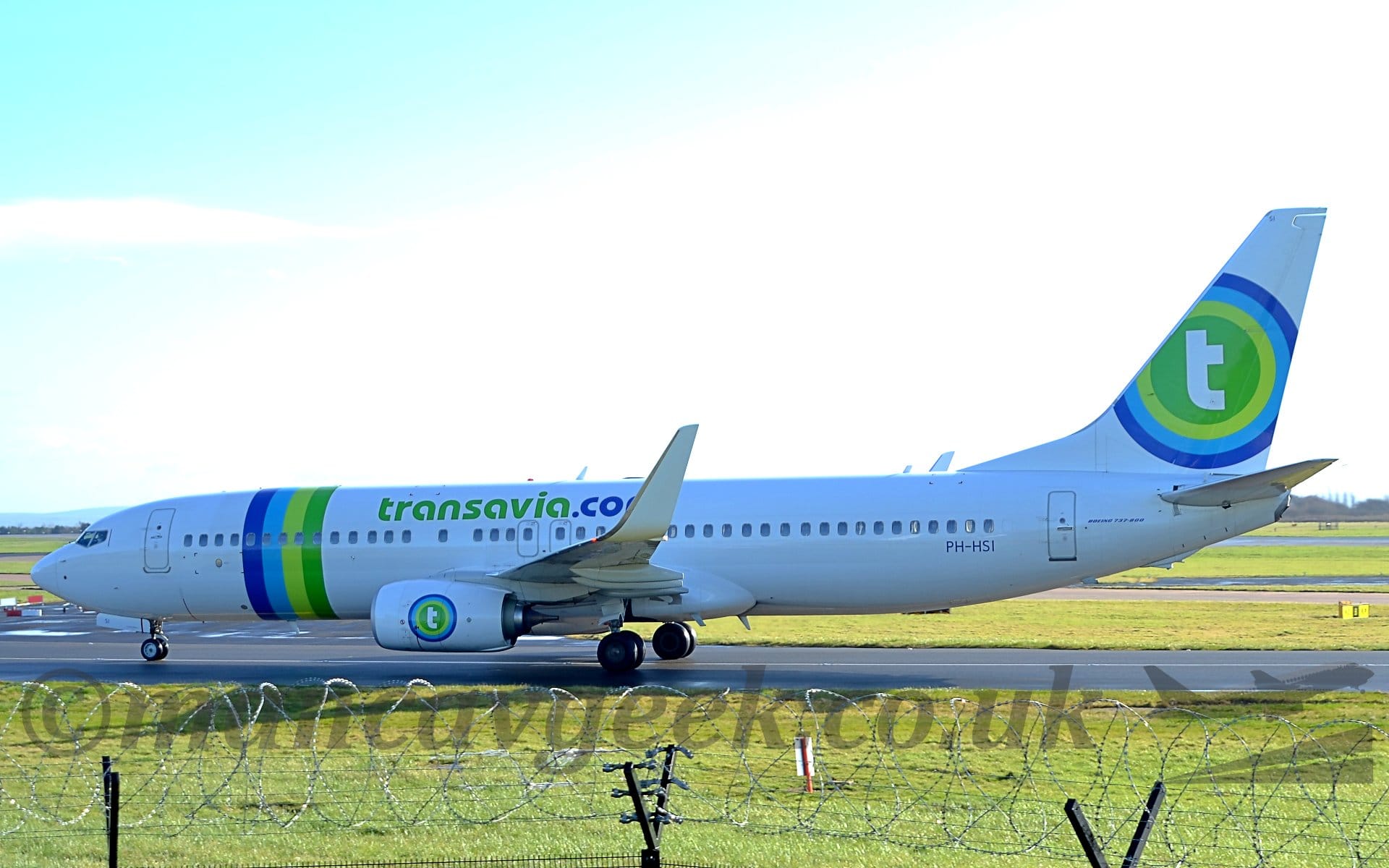 Side view of a white, twin engined jet airliner taxiing from right to left. The plane has purple, light blue, lime green, and dark green bands around the forward fuselage, and green and purple "Transavia[dot]com" titles on the upper fuselage. There is a dark green circle on the tail with a white lower-case letter "t" in the centre, the circle surrounded by concentric light green, light blue, and purple rings. The same logo is repeated on the sides of the engine pods under the wings. Green grass fills the foreground, with more grass leading off into the distance, while bright but hazy sky fills the rest of the frame, with patches of blue in the top left corner.