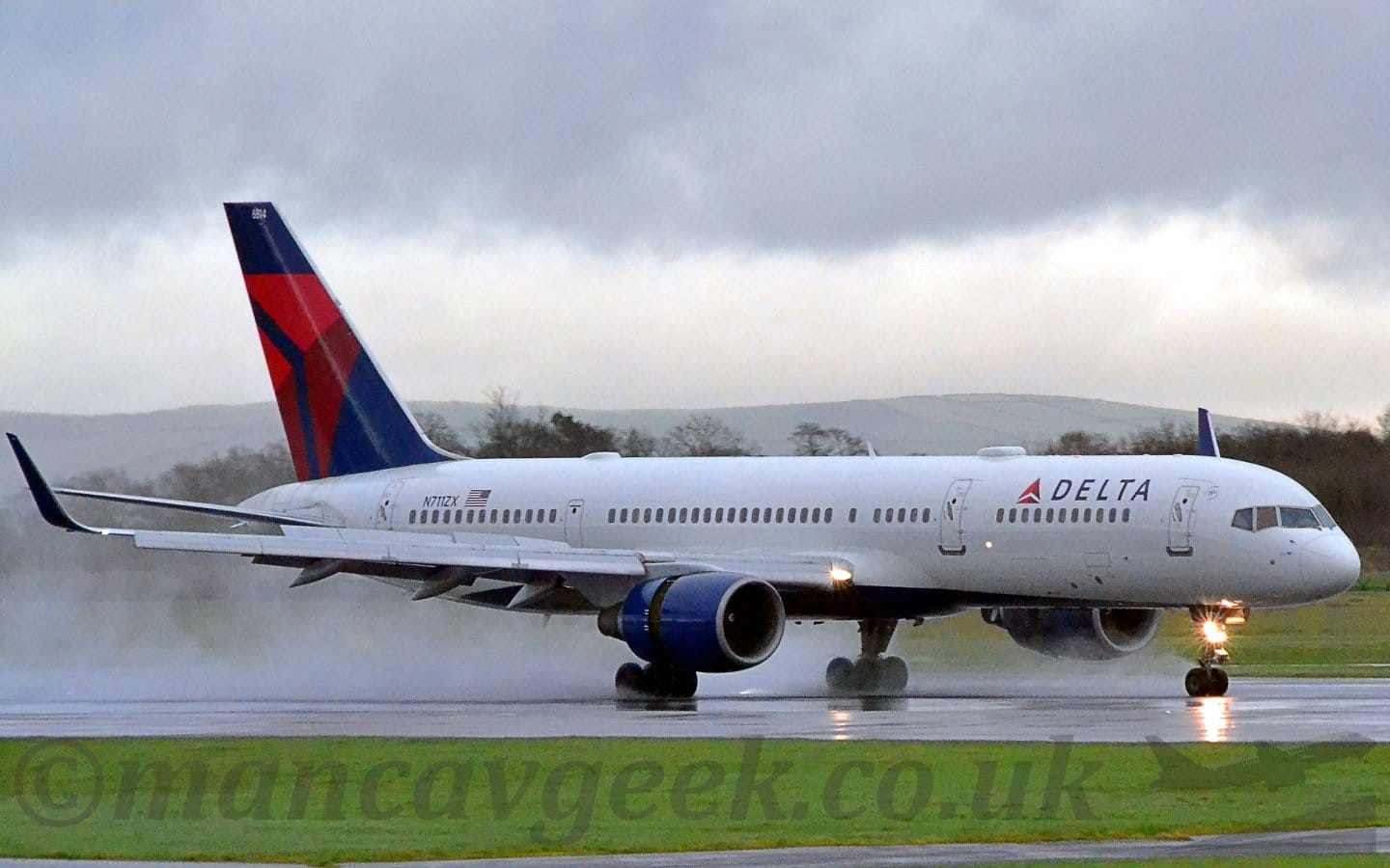 Side view of a white, twin engined jet airliner slowing down as it moves from left to right along a wet, black runway. Spray is being kicked up by the wheels and the exhaust from the engines. The plane has black "Delta" titles on the upper forward fuselage, with the registration "N711ZX" on the upper rear fuselage, next to an American flag. The engine pods slung under the wings have the rear section pushed back, allowing the thrust reversers to push air forwards to slow the plane down. There is a bright light mounted on the nose wheel, pointing forwards. Green grass fills the bottom of the frame in the foreground, with more grass on the other side of the runway, leading up to trees in the distance. In the far distance, rolling hills give way to stormy grey clouds.