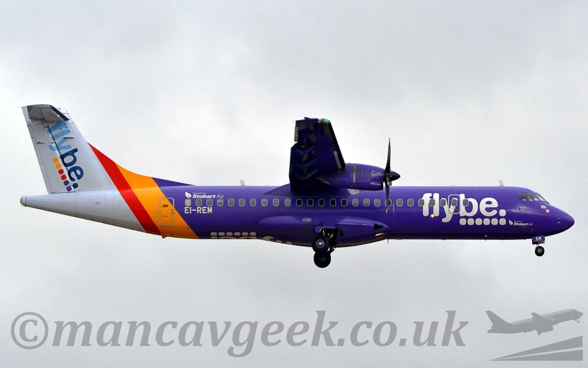 Side view of a high-winged, twin propellor-engined purple airliner flying from left to right at a very low altitude, with undercarriage extended and flaps deployed from the rear of the wings, suggesting it is about to land. The plane is mostly a lush purple, with a yellow and red band around the rear fuselage, and a whitetail and tail-cone. There are large white "FlyBe" titles over 6 white dots on the forward fuselage, a design replicated diagonally on the tail, only this time the text is in 2 shades of blue, and with 2 yellow dots, 2 red dots, and 2 purple. There are white "Stobart Air" titles under the forward fuselage, just over the nose-wheel doors, repeated over the rear passenger cabin windows, and the registration "EI-REM" just under those windows, also in white.