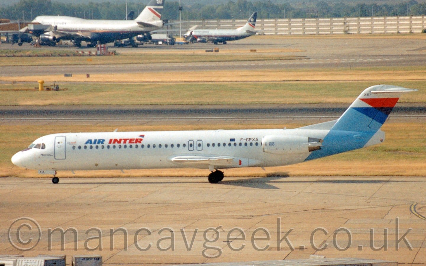 Side view of a white, twin engined jet airliner with the engines mounted on the sides of the rear fuselage, taxiing from right to left. The plane has blue and red "Air Inter" titles on the upper forward fuselage, and a light blue band wrapping around the rear fuselage and up into the tail, topped by overlapping dark blue and red triangles. Grey concrete apron fills the foreground, with strips of grass in the background delineating runways and taxiways. In the distance, a couple of grey and dark blue, almost black, jet airliners can be seen on the left of the frame, with a tall concrete wall behind them, and trees beyond that.