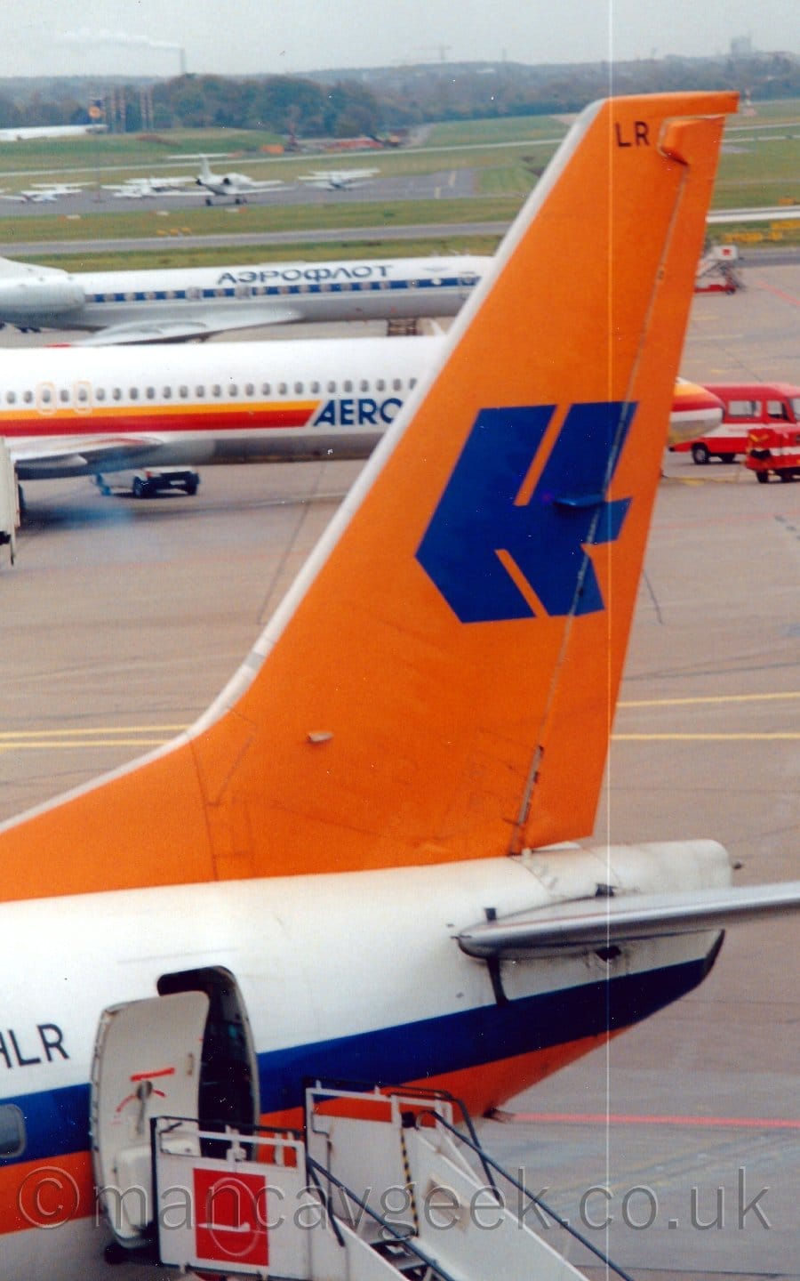 Closeup of the white rear fuselage and bright orange tail of a jet airliner parked facing to the left. There is an orange and blue stripe running along the body, and  a stylised upper-case letter "H" in blue on the orange tail. A set of white mobile airstairs is connected to the open rear door. A pair of white jet airliners can be seen in the background on the left of the frame, with light aircraft and bizjets parked on an apron surrounded by grass further back, under cloudy grey sky.