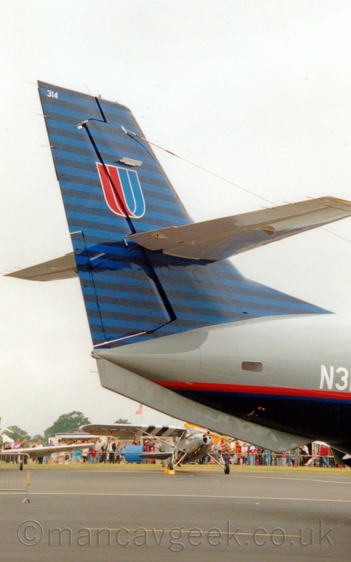 Closeup of the blue and black striped tail of a grey and blue airliner parked facing to the right. There is a red and blue stylised letter "U" on the tail, and a thin red, orange, and blue stripe running along the body. In the background, under the planes rear fuselage, a couple of single engined light aircraft can be seen, with crowds of people behind them. Dreary grey, cloudy skies fill the rest of the frame.