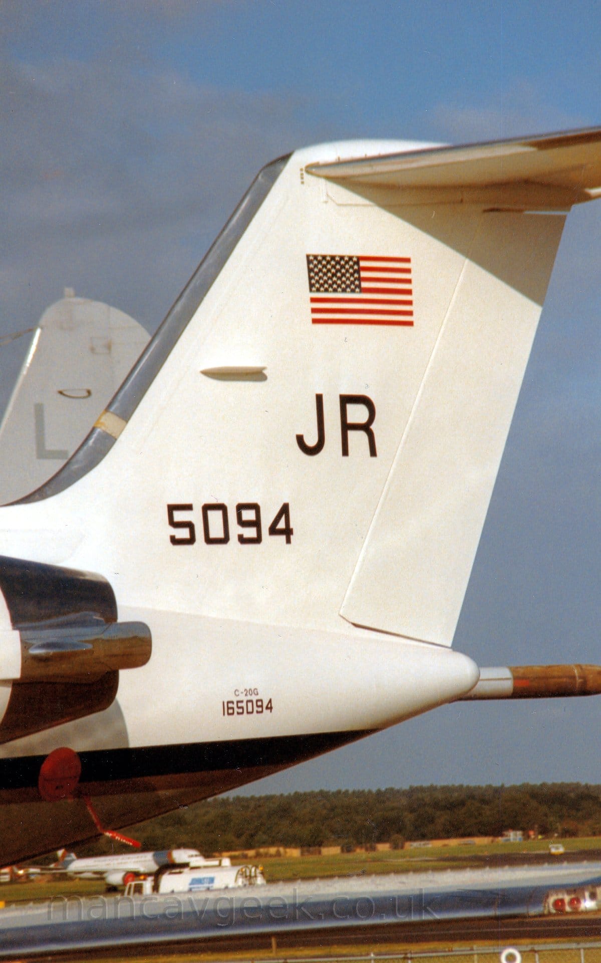 Closeup of the white tail and rear fuselage of a twin engined BizJet with the rear of the engines visible mounted on the sides of the rear fuselage, parked facing to the left. There is a US flag at the top of the tail, with large letters "JR" in black underneath, and the number "5094" below that and very slightly further forward. The tailcone has text "C-20G" in very small letters, with the number "165094" in slightly larger letters below that. A black stripe runs along the body, wrapping around under the tailcone. The planes wing stretches across the bottom of the frame, with a white, twin engined jet airliner visible over the top, with trees filling the skyline , under blue-grey skies.