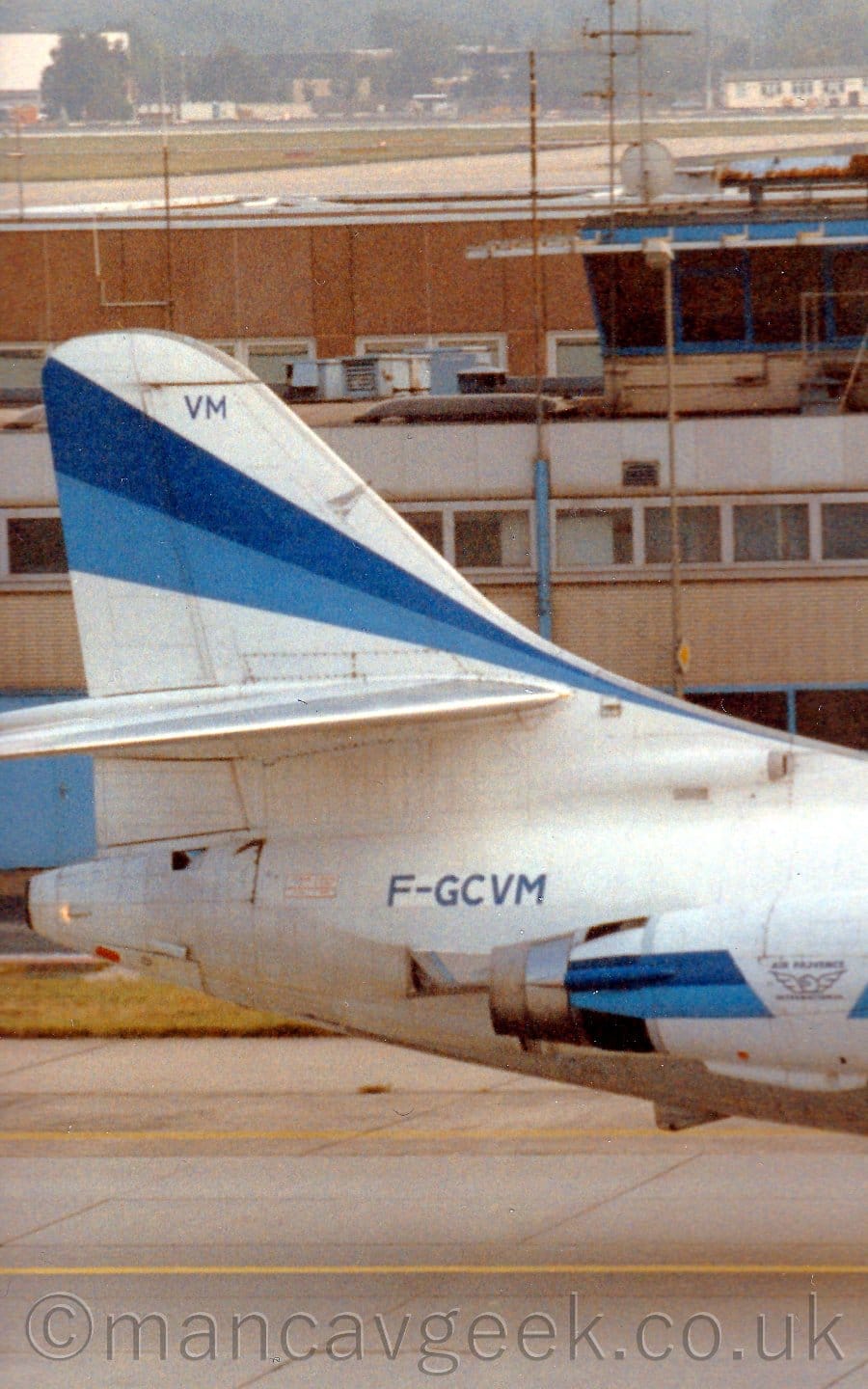 Close up of the white rear fuselage and tail of a twin engined jet airliner facing to the right with the engines mounted on the sides of the rear fuselage. The tail has a two-tone blue running from the lower front edge to the top rear, getting wider as it goes. A similar stripe runs along the engine pods, with a white section in the middle with a winged logo with "Air Provence" title above it. The registration "F-GCVM" is on the upper rear fuselage, just aft of the engines, in blue. A wide building in shades of brown stretches across the background, with trees and low buildings in the distance.