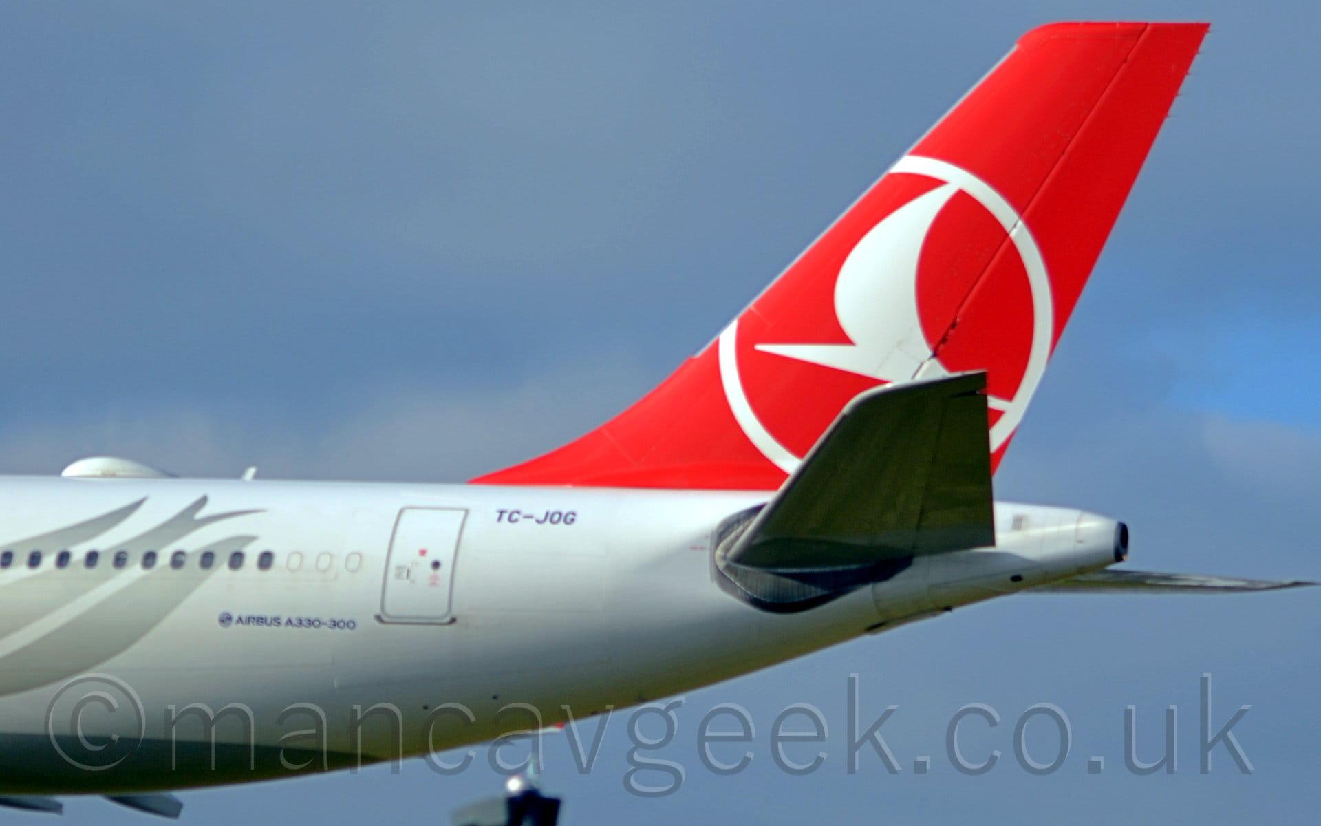 Closeup of the white rear fuselage and red tail of a jet airliner flying from right to left at a very low altitude. The rear fuselage has a silver flower head on the rear fuselage, while the red tail has a stylised white flying bird inside the outline of a white circle. Blue sky with hazy grey cloud fills the rest of the frame.