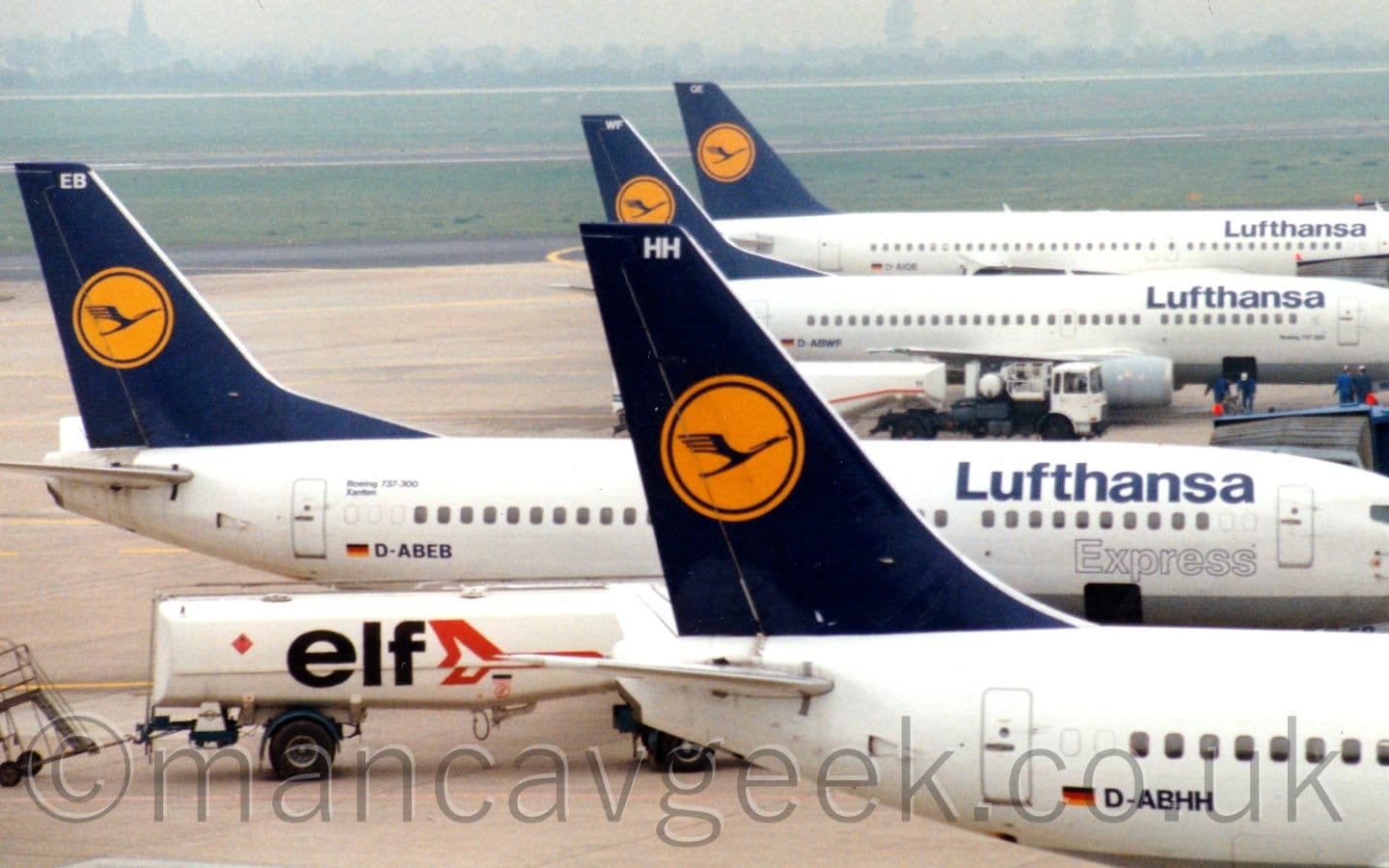 The tails of four white jet airliner with blue tails lined up facing to the right at a slightly misty airport, the 2nd in line parked further to the left. The planes all have dark blue tails, with yellow circles containing a blue stylised flying bird in the middle. They all also have blue "Lufthansa" titles on the upper forward fuselage. There is a white fuel tanker parked between the first 2 planes. Green grass in the distance fills the top of the frame, leading up to trees on the horizon, under grey skies.